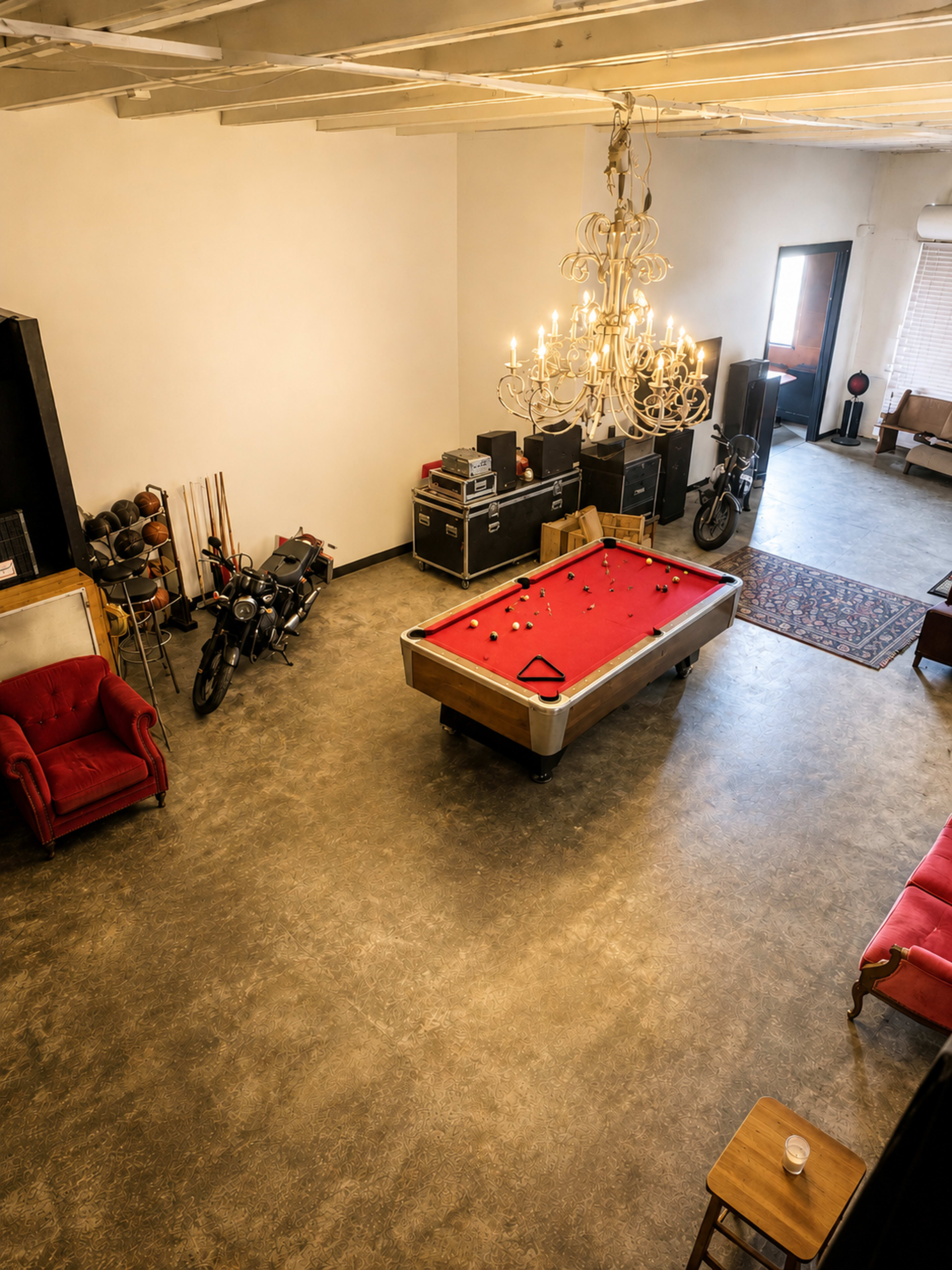 The image shows a spacious loft with a red pool table at the center, surrounded by motorcycles, vintage furniture, and a chandelier hanging from the ceiling.