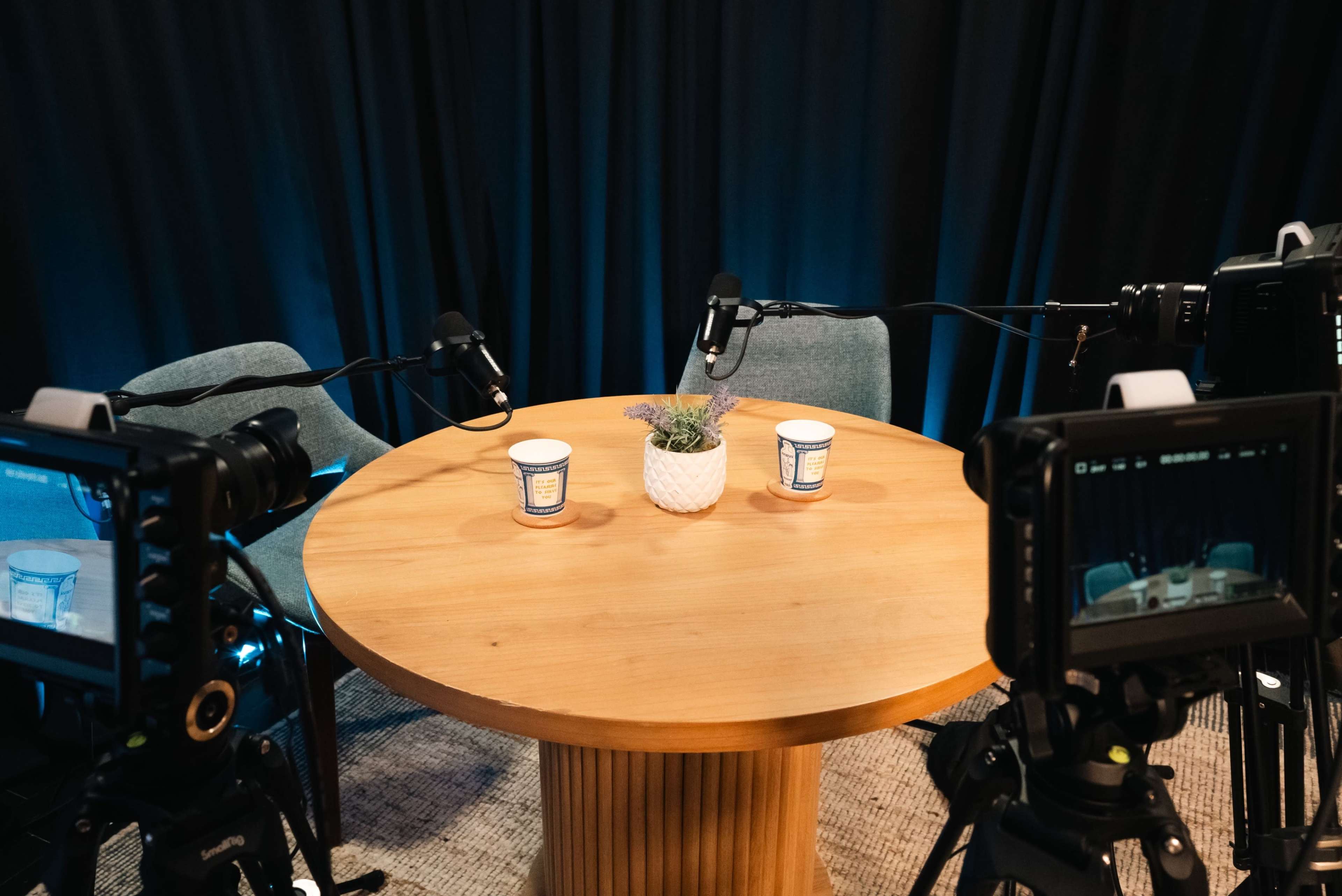 A round wooden table with two cups and a small plant is set between two chairs, surrounded by cameras and microphones.