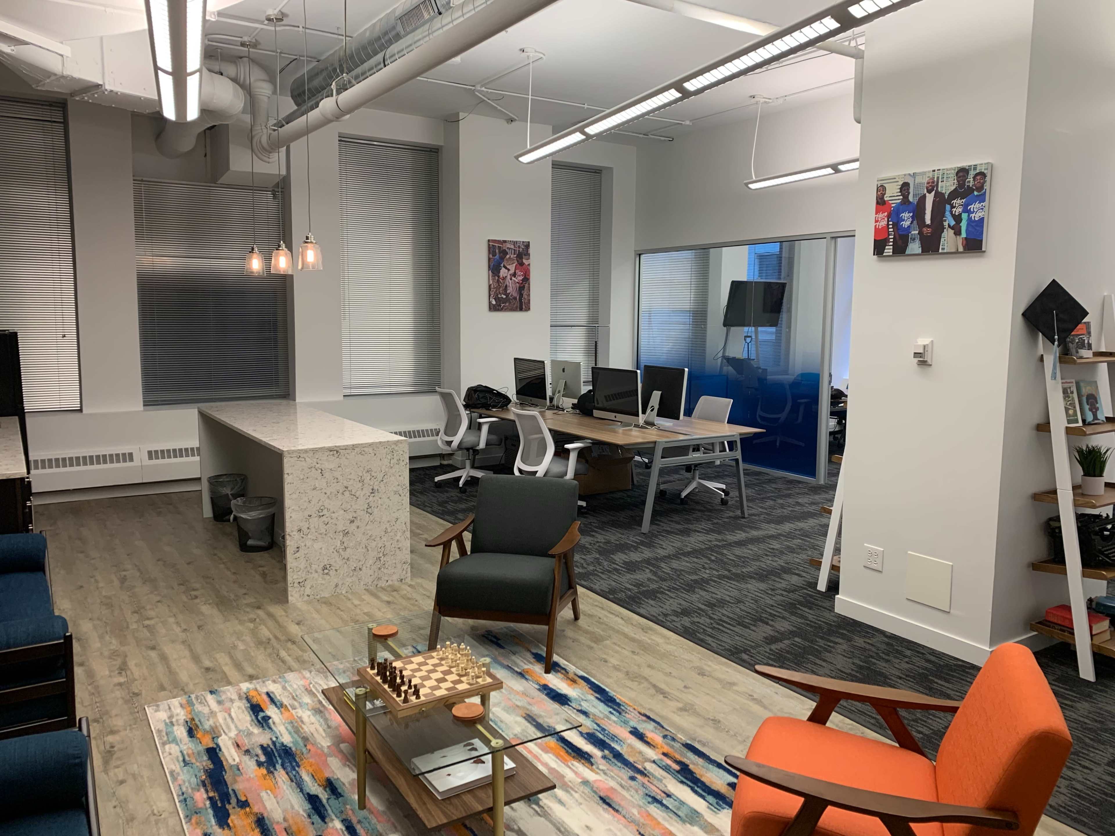 A modern office space featuring a seating area with chairs, a communal table, and multiple workstations in a well-lit environment.