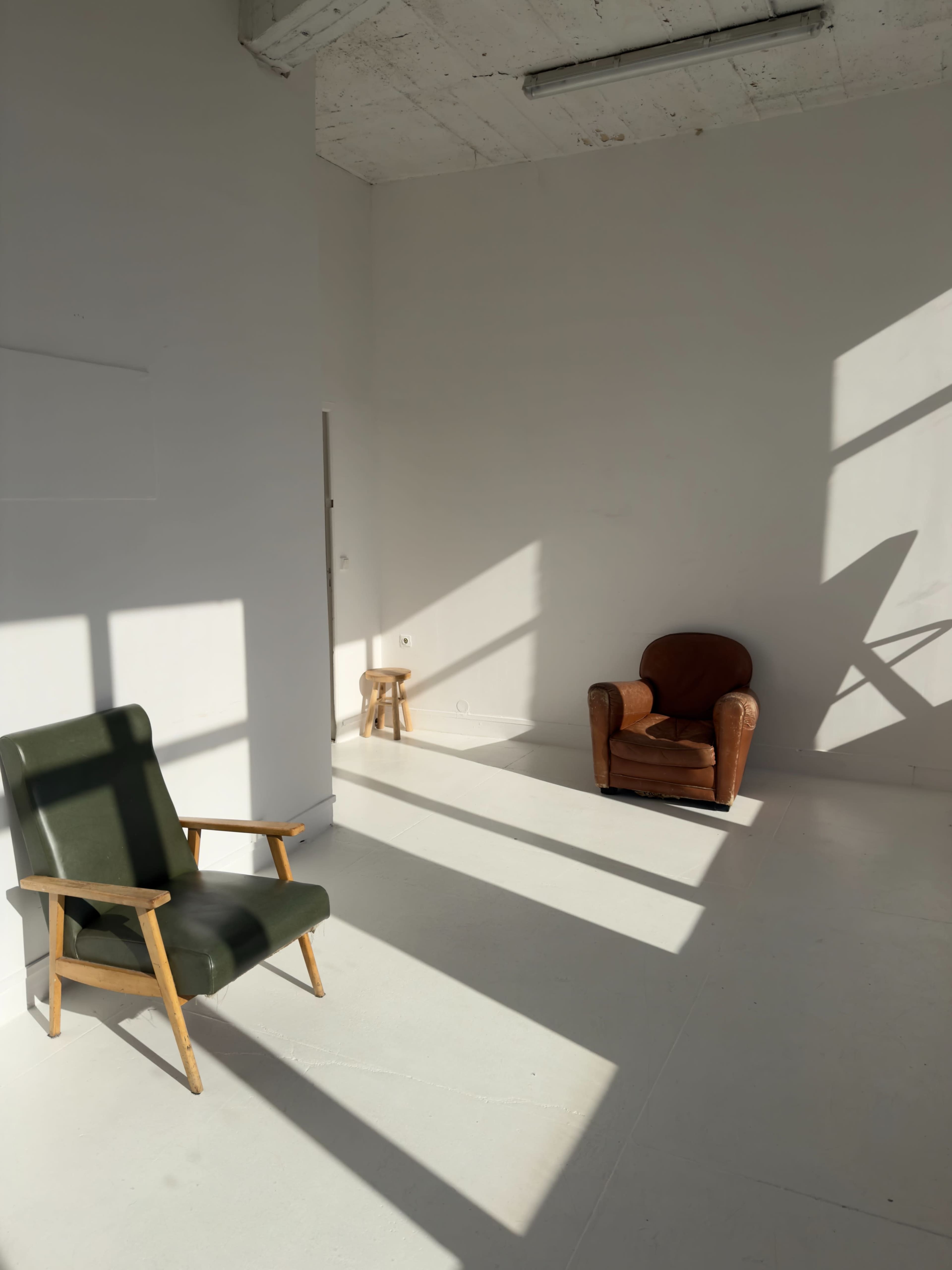 The image shows a minimalistic room with two distinct chairs, one green and wooden and the other brown and leather, illuminated by diagonal sunlight casting shadows on the walls.