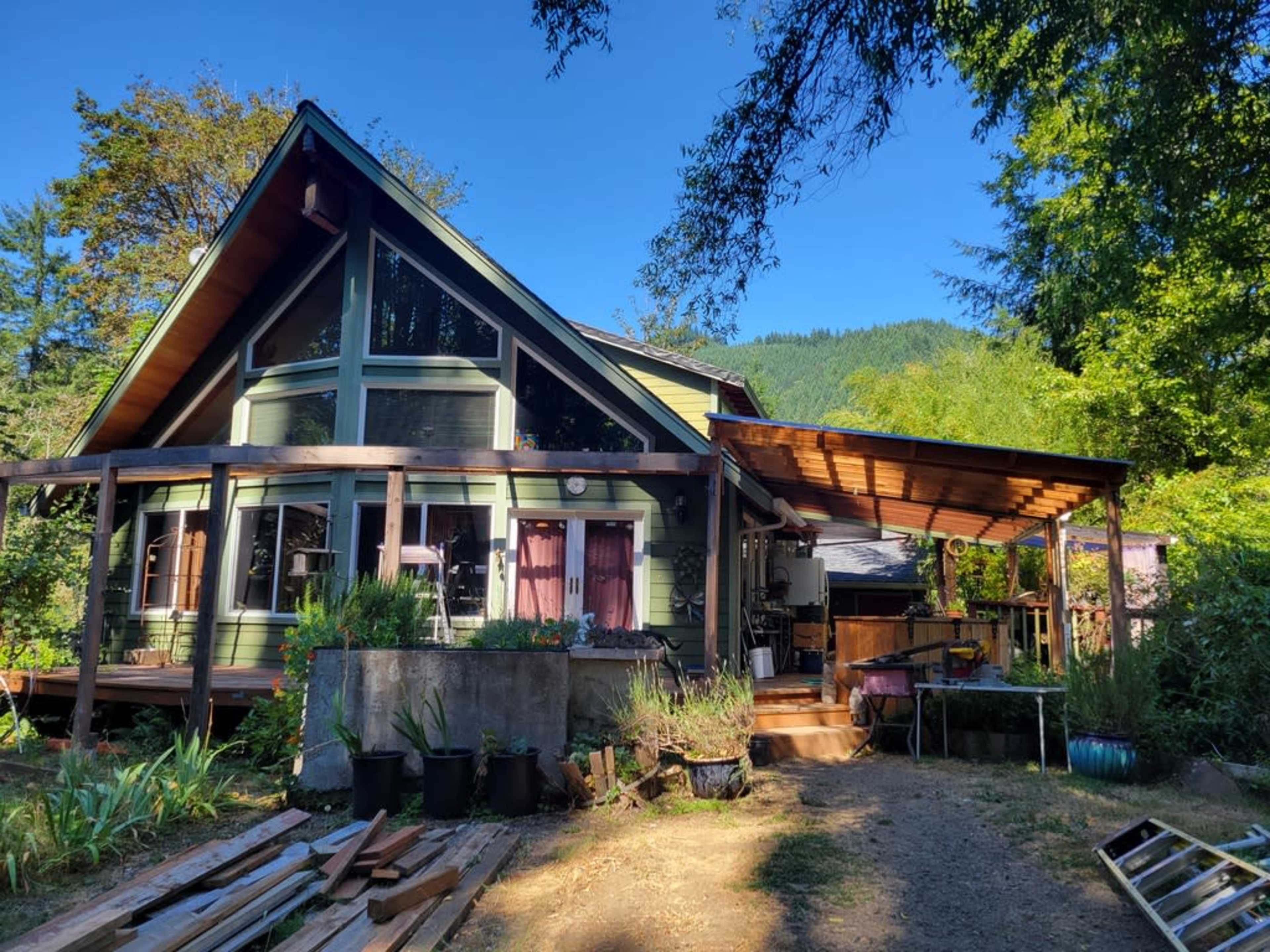 A green and brown house with a large deck, surrounded by trees and gardening supplies.