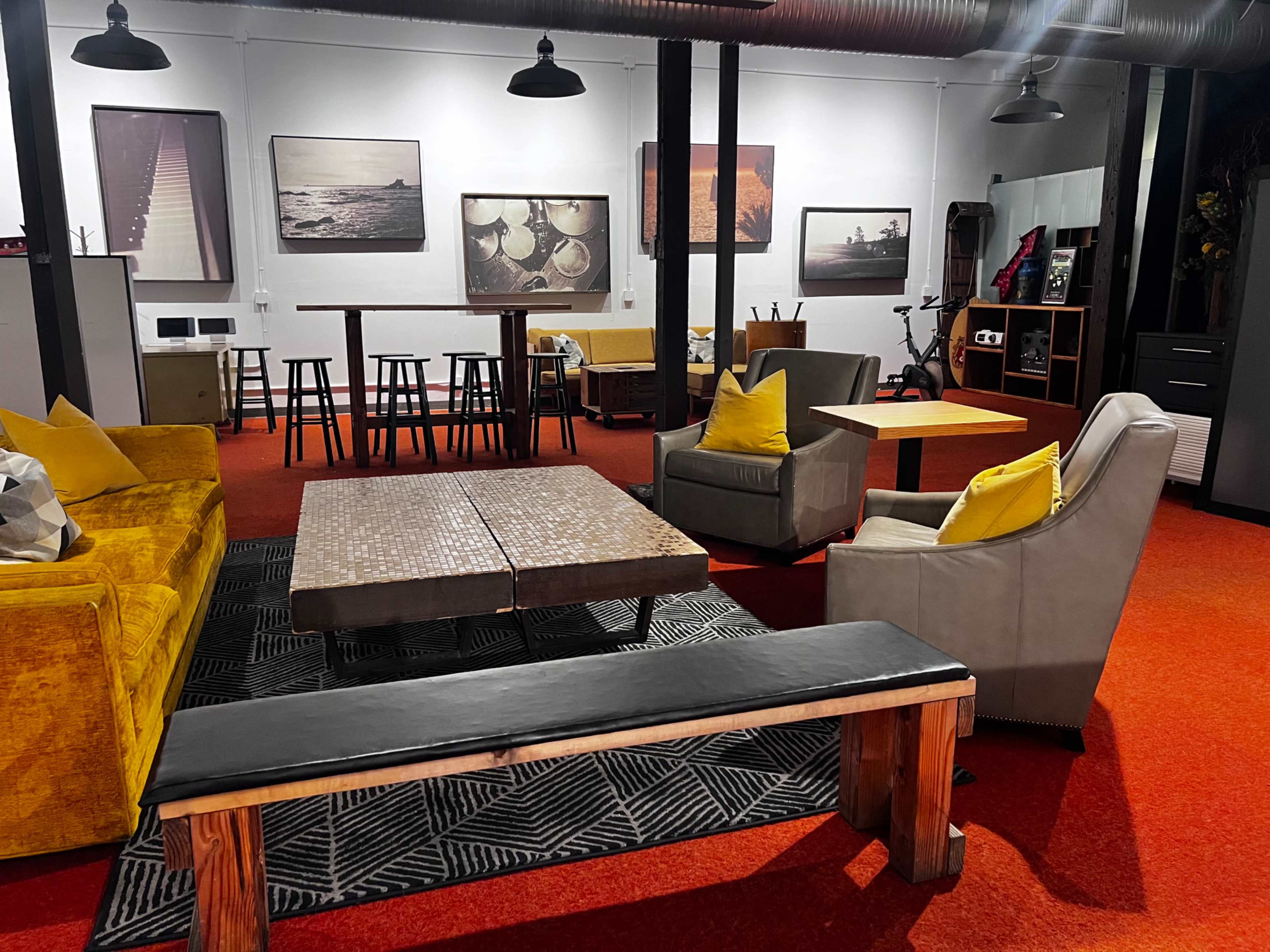 The image shows a modern lounge area featuring colorful seating, a coffee table, and abstract art on the walls, with bright orange carpeting.