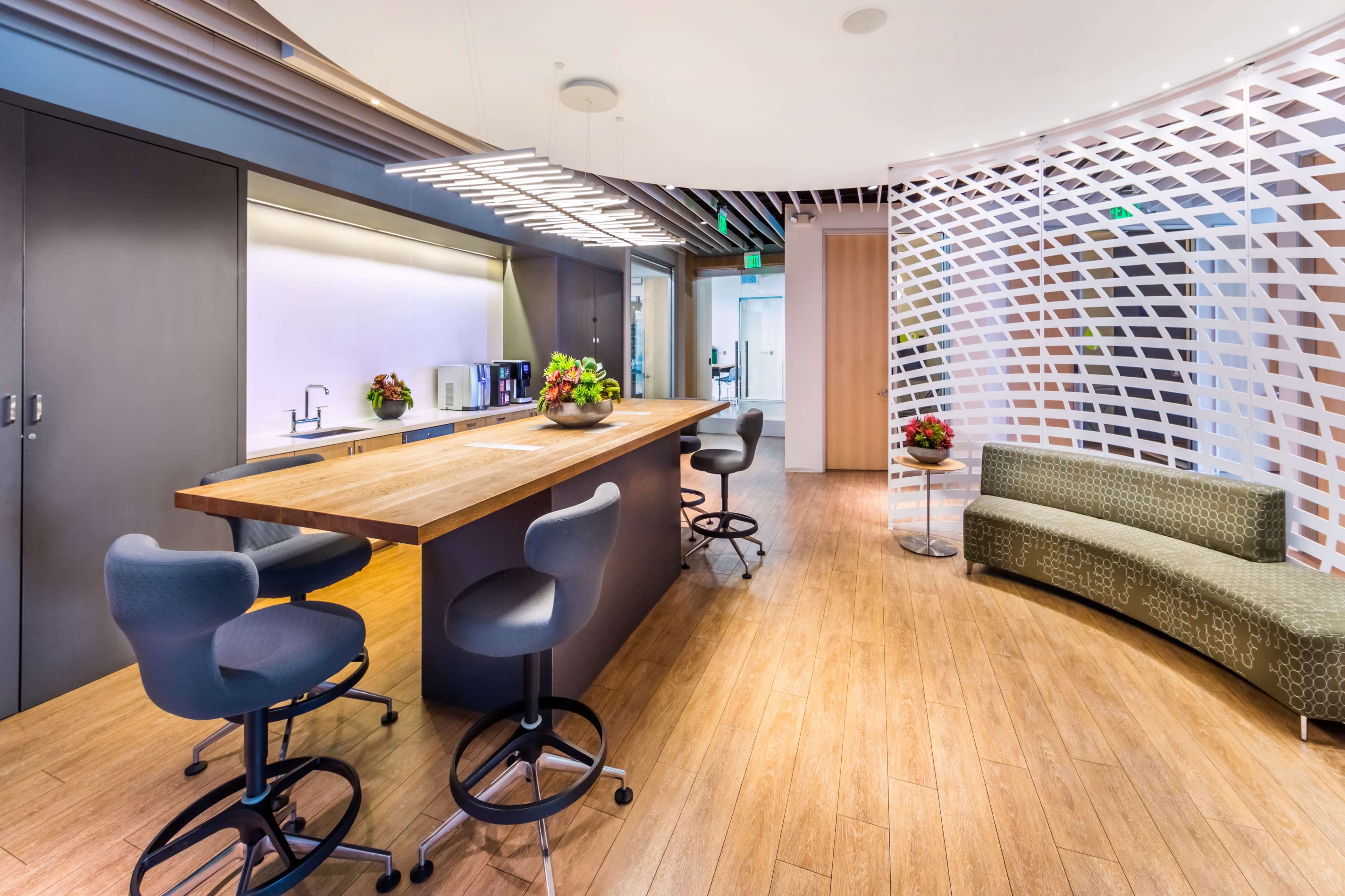 A modern kitchen area with a wooden bar top, high stools, and a curved sofa, surrounded by light-colored walls and decorative elements.