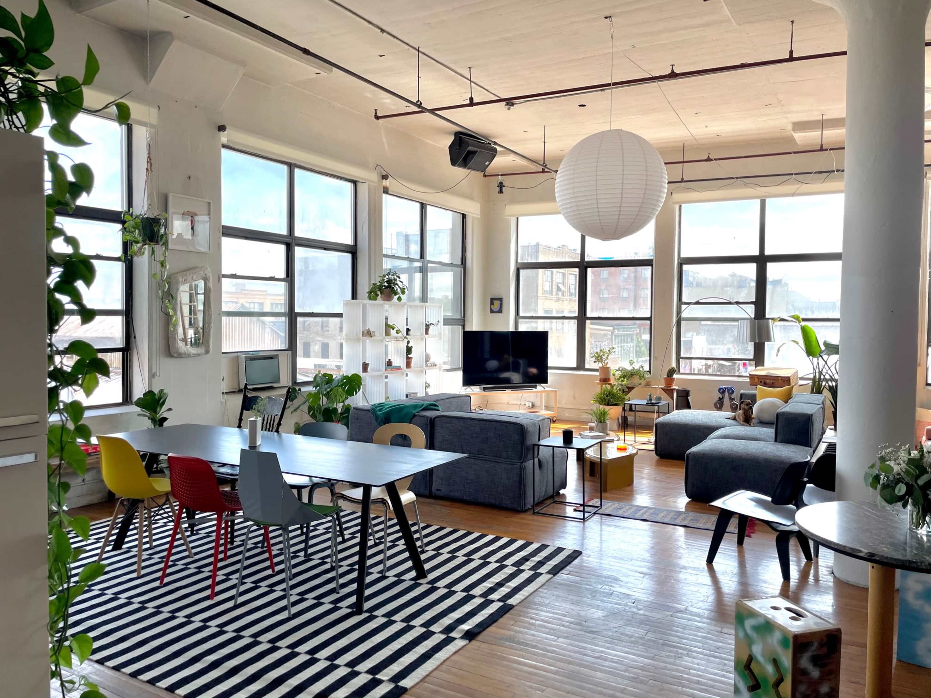 The image shows a bright, open living space with a large window, featuring a mix of seating areas, a dining table, and decorative plants.
