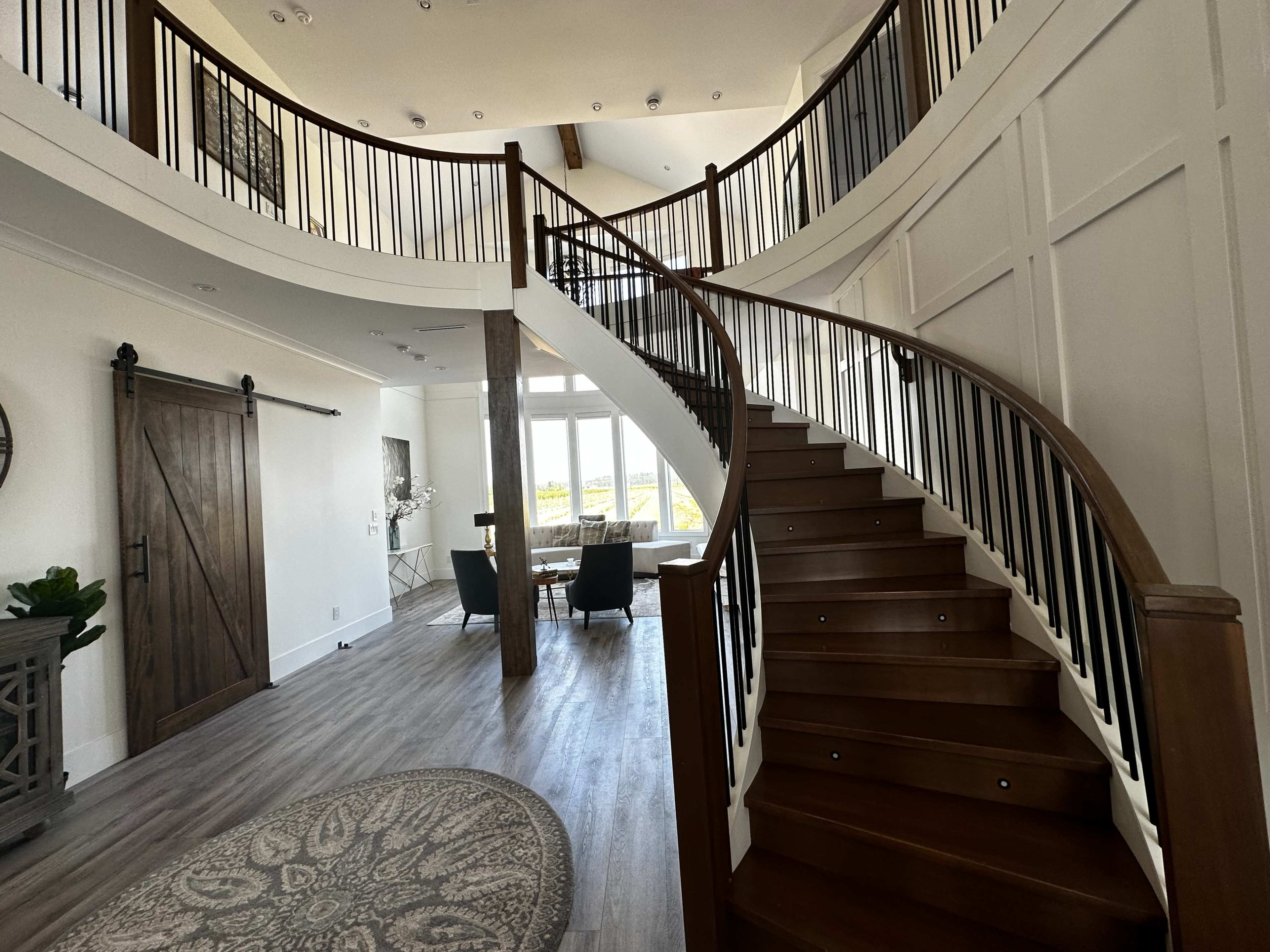 A spacious foyer featuring a curved wooden staircase, a large round rug, and a dining area with large windows.