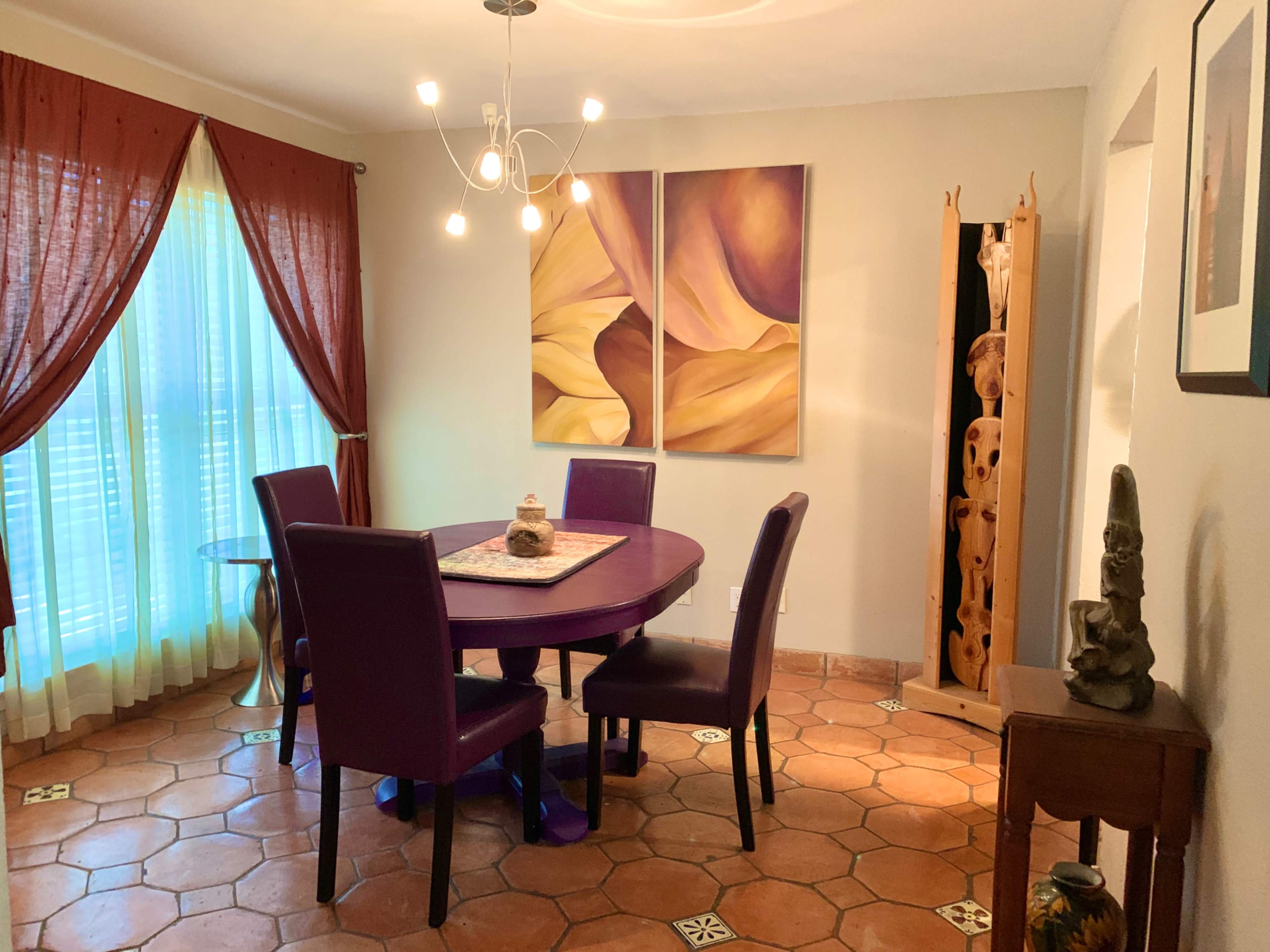A dining area features a round purple table surrounded by four matching chairs, with large windows dressed in sheer curtains and two abstract paintings on the wall.