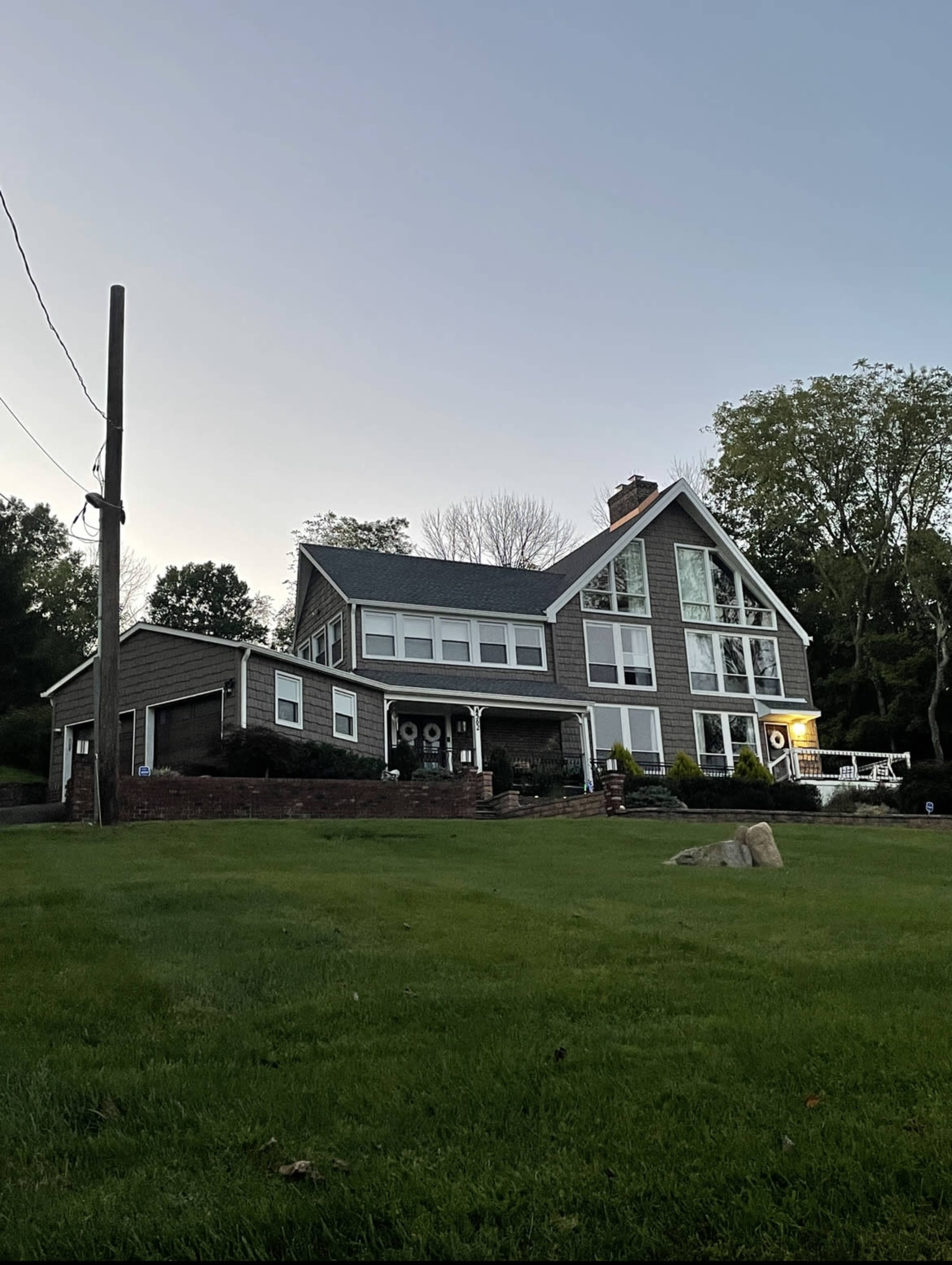 A large, modern house with multiple windows and a sloped roof is set on a grassy lawn bordered by a power pole and trees.