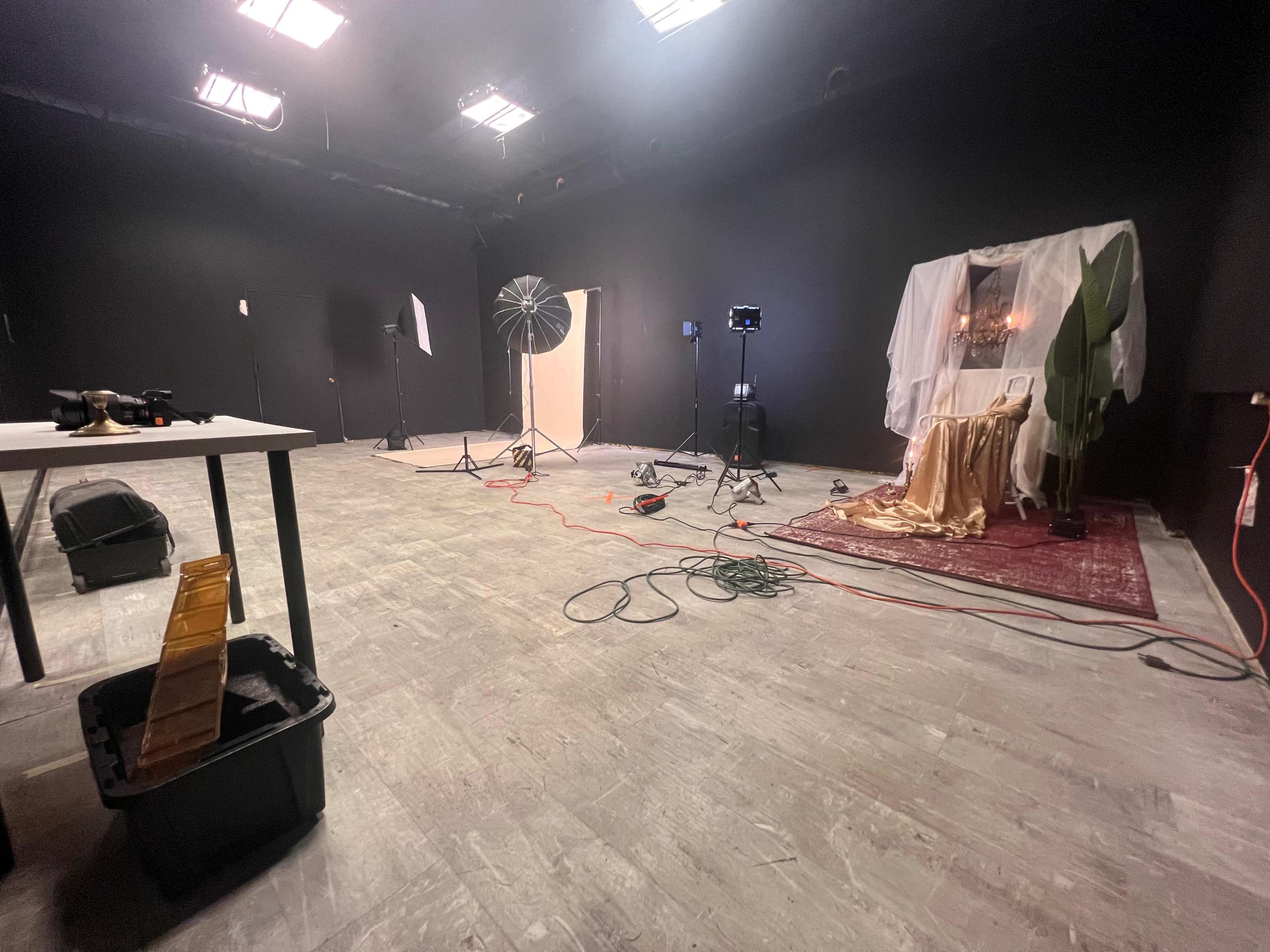 The image shows a dimly lit studio space with black walls, featuring a setup for a photo shoot, including lights, a backdrop, and various equipment scattered around.