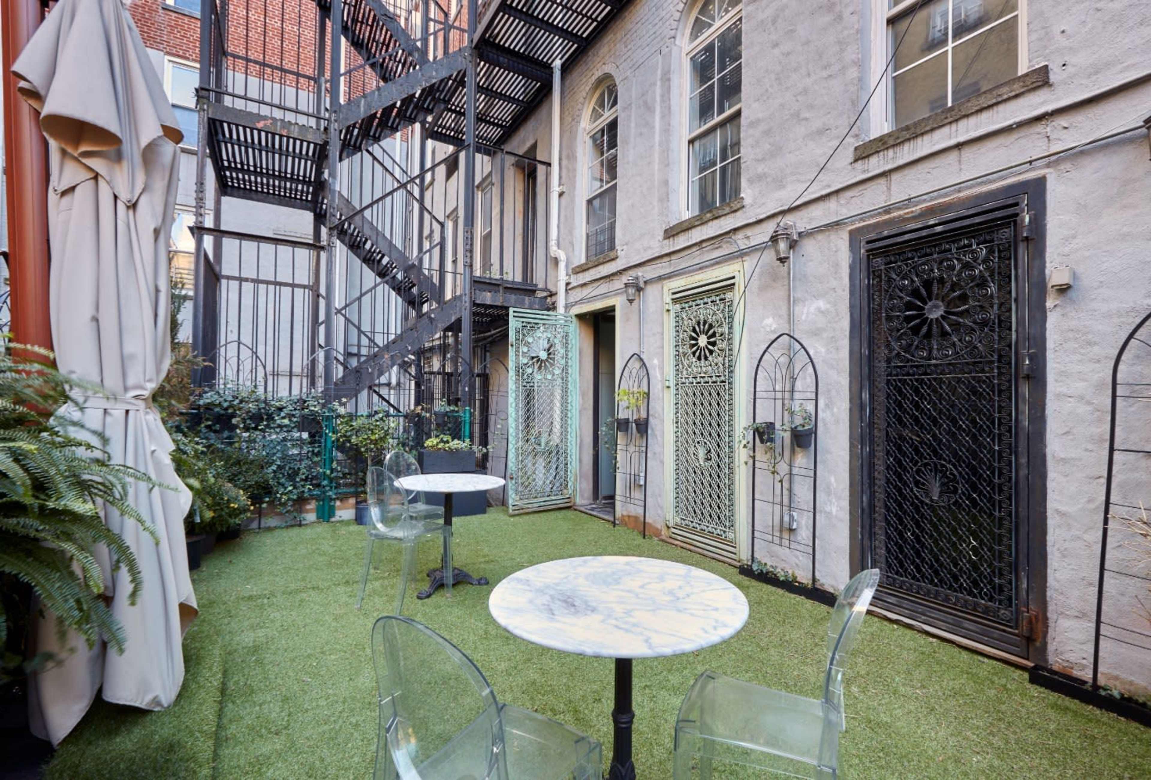 A small courtyard with artificial grass, two marble-topped tables, and a staircase leading to upper levels, surrounded by buildings with distinct doors.