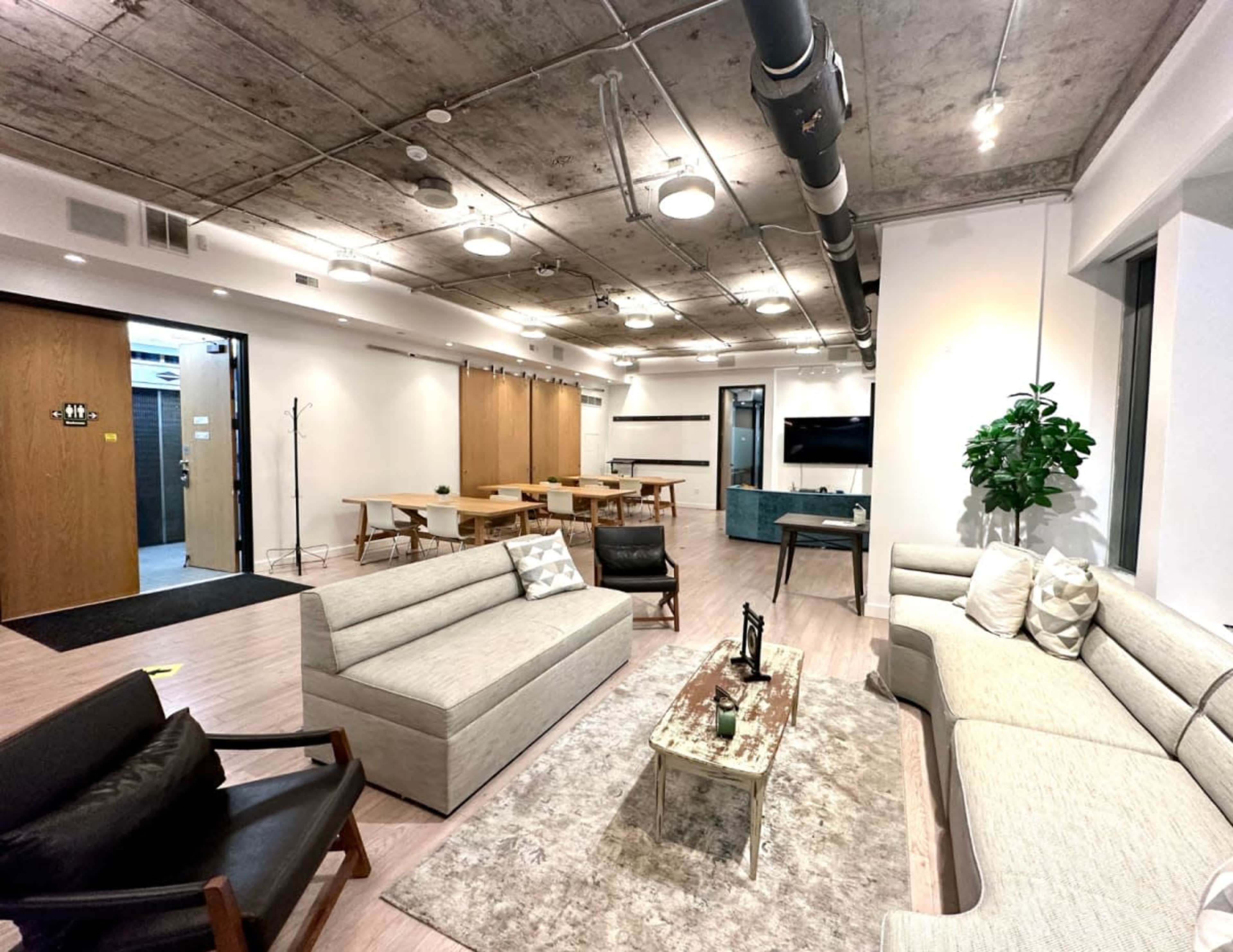 The image shows a modern, open office lounge with sofas, a coffee table, and tables set up for meetings, against a backdrop of concrete ceilings and large windows.