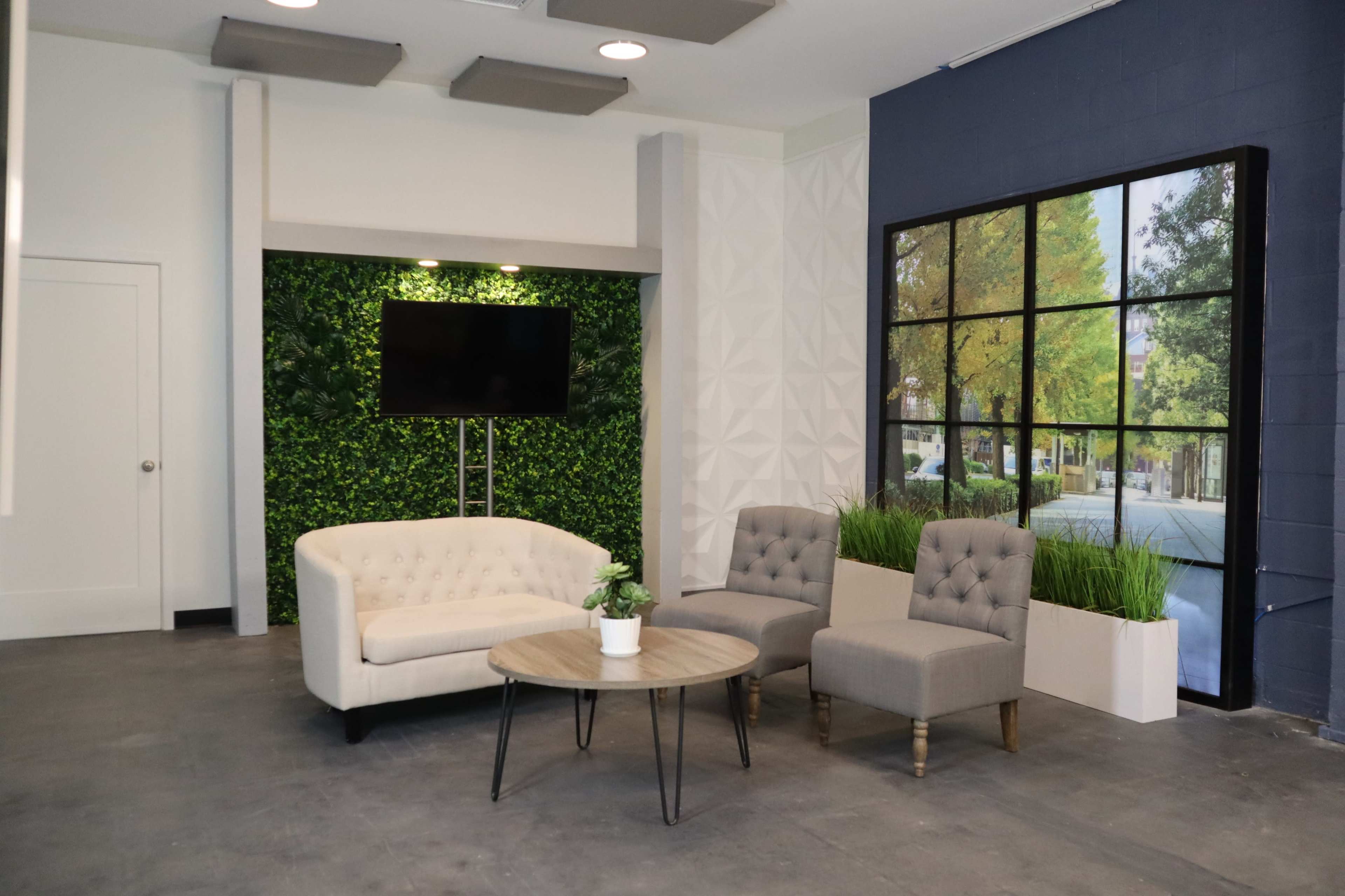A modern lounge area with a white sofa, two gray chairs, a round wooden table, a television on a wall, and a large window graphic depicting a tree-lined street.