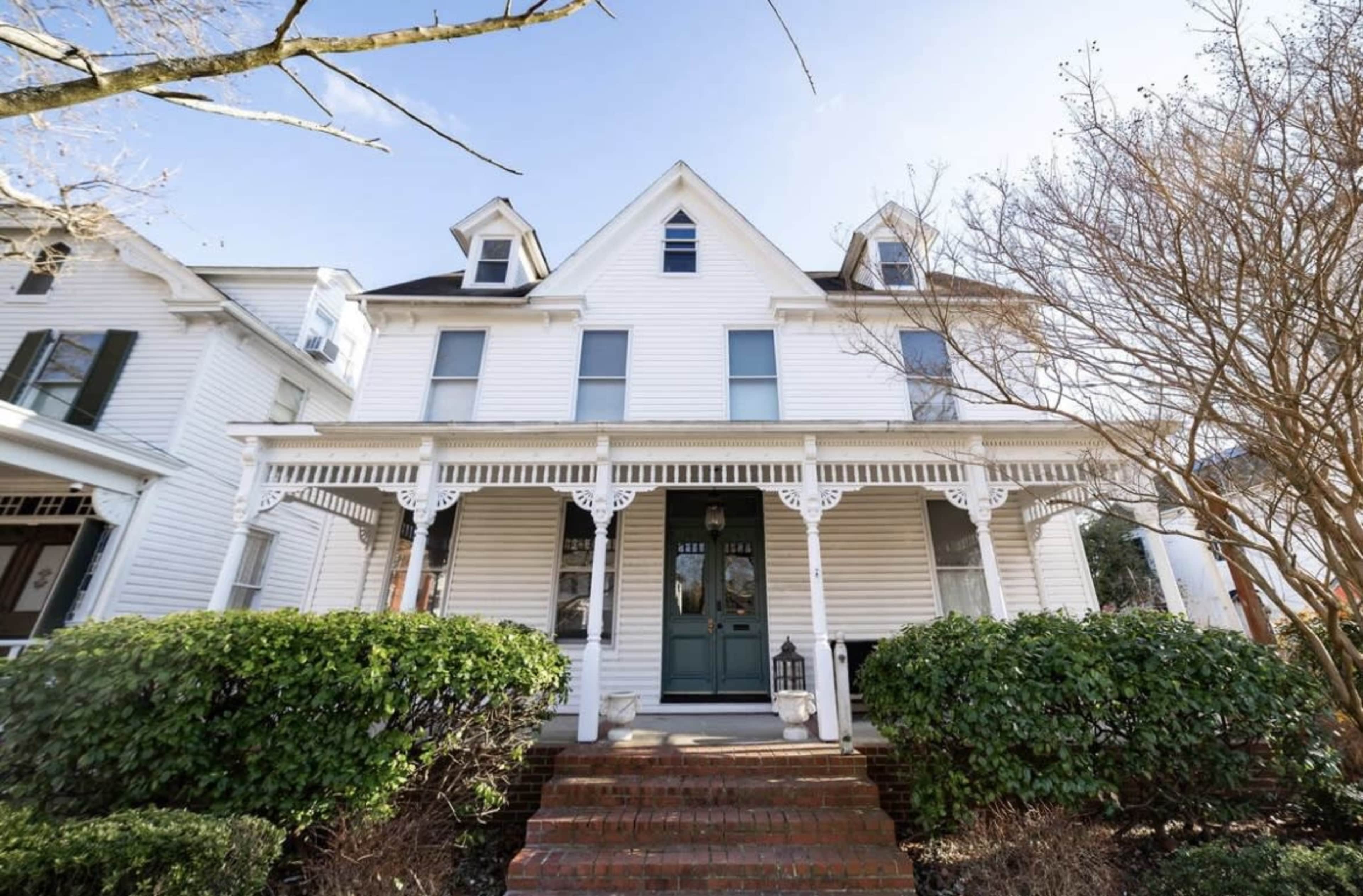 The image shows a Victorian-style house with a white exterior, a front porch supported by decorative columns, and manicured bushes surrounding the entrance.