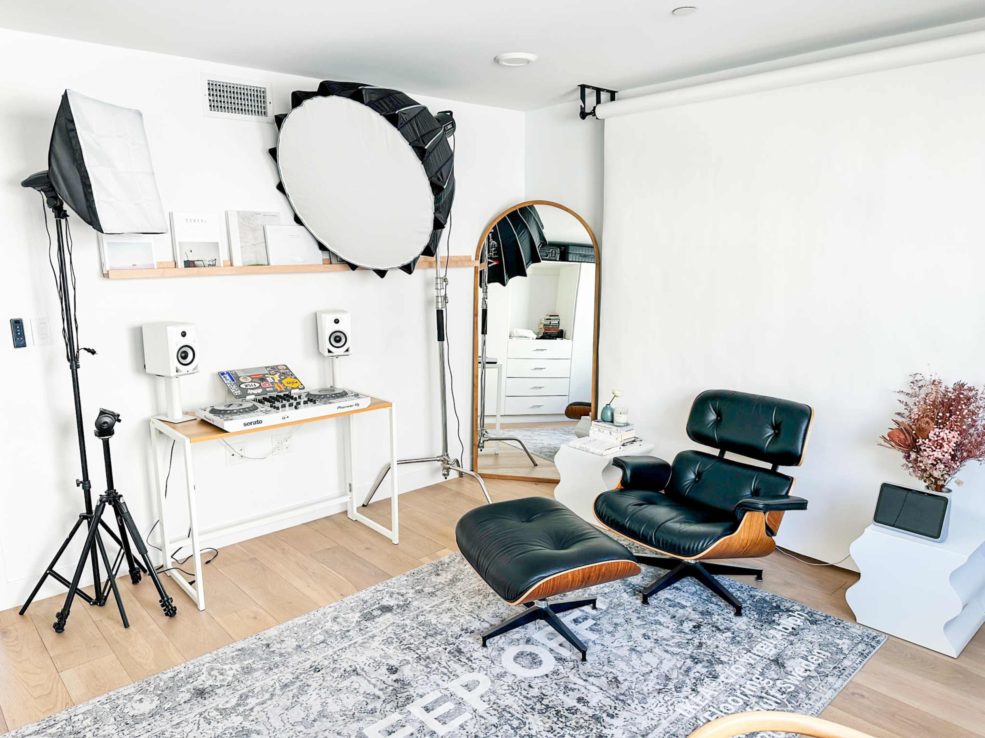 The image shows a well-organized room featuring a black leather chair, a mirrored dresser, studio lighting equipment, and a small table with audio equipment.