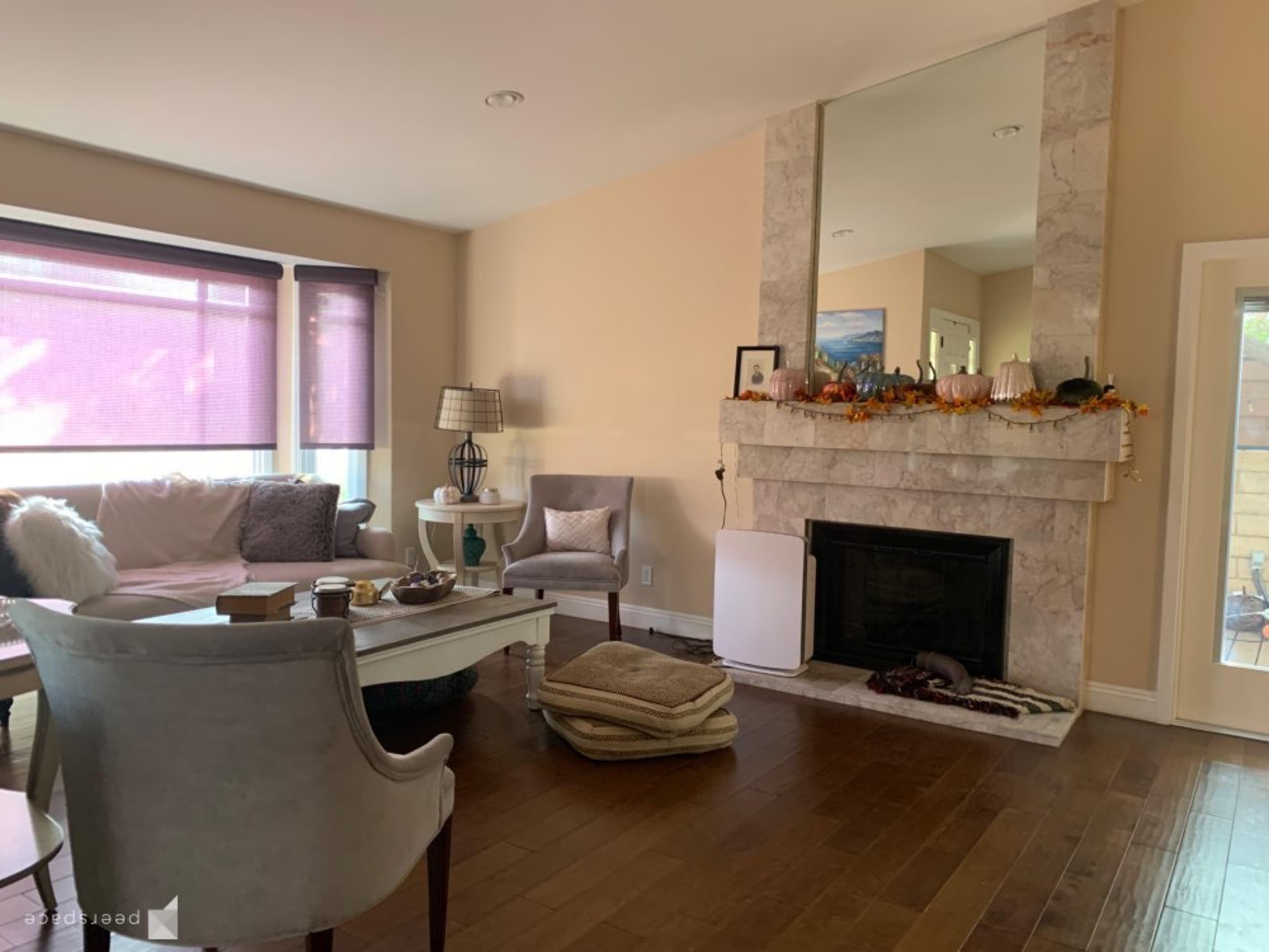 A living room with a stone fireplace, a large mirror above it, and a light-colored sofa arranged around a coffee table on wooden flooring.