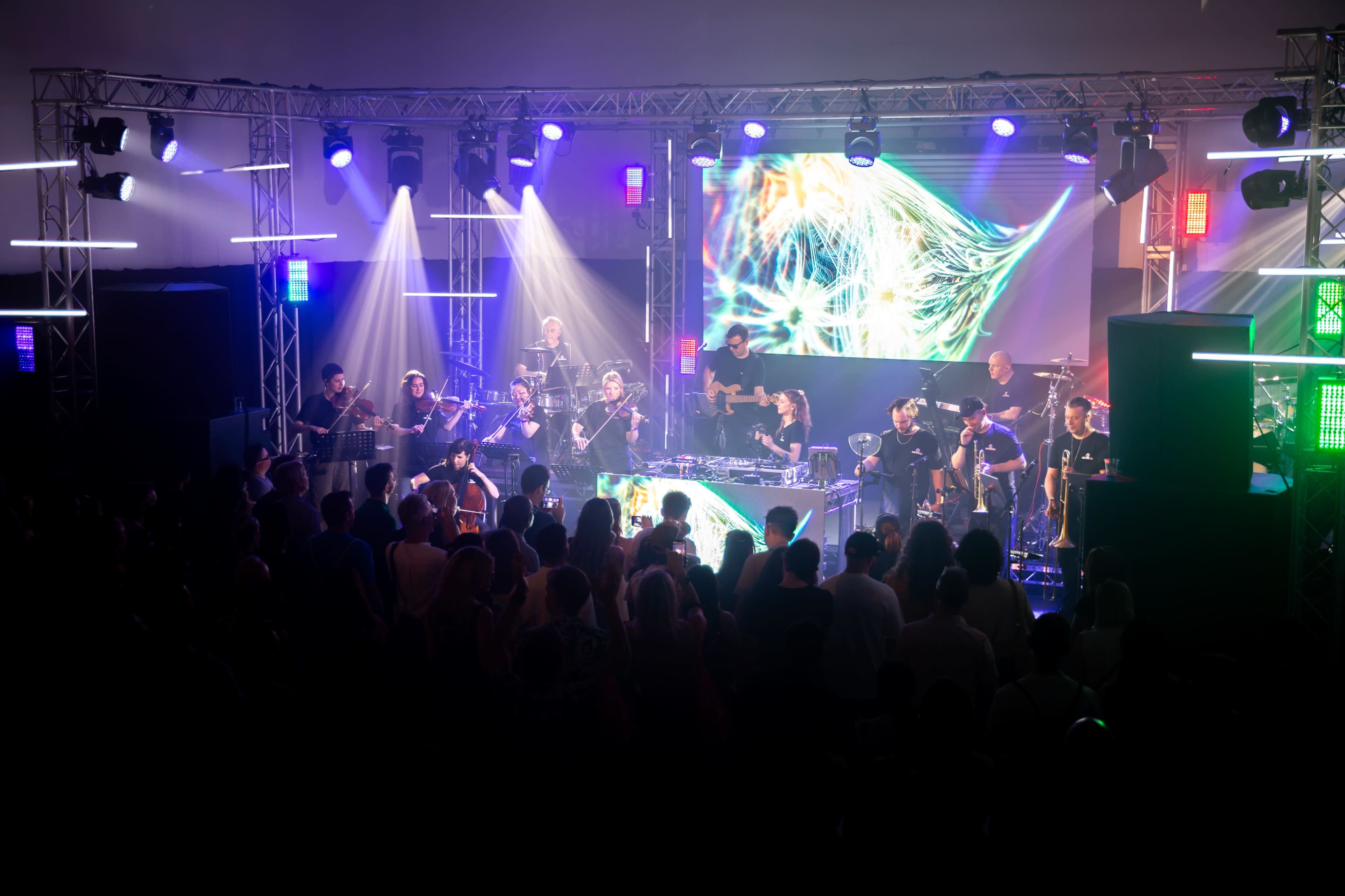 A large crowd enjoys a live music performance by a band illuminated by colorful stage lights and visual effects.