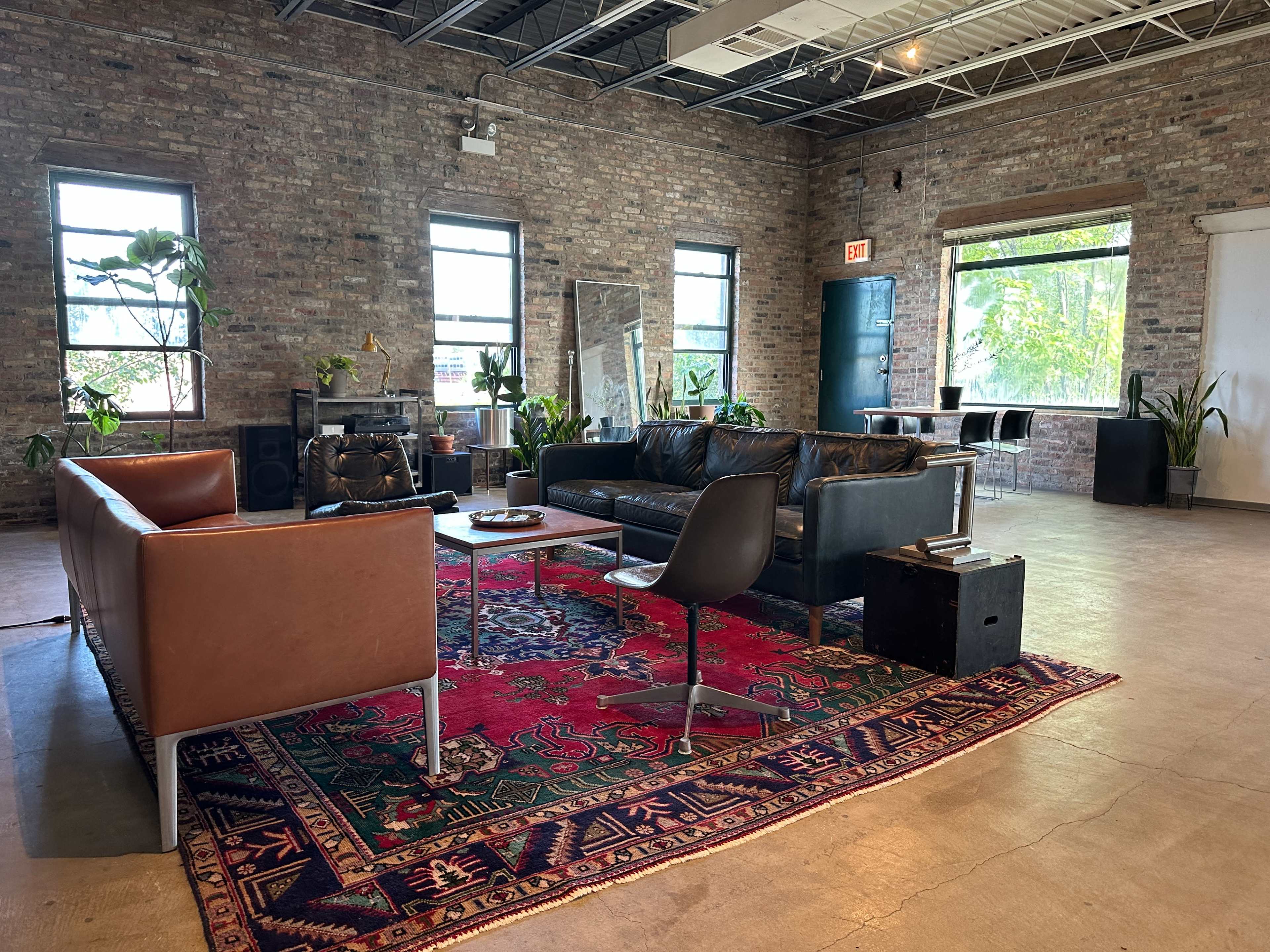 The image shows a spacious industrial-style living area with two sofas, a coffee table, and large windows allowing natural light, surrounded by plants and brick walls.