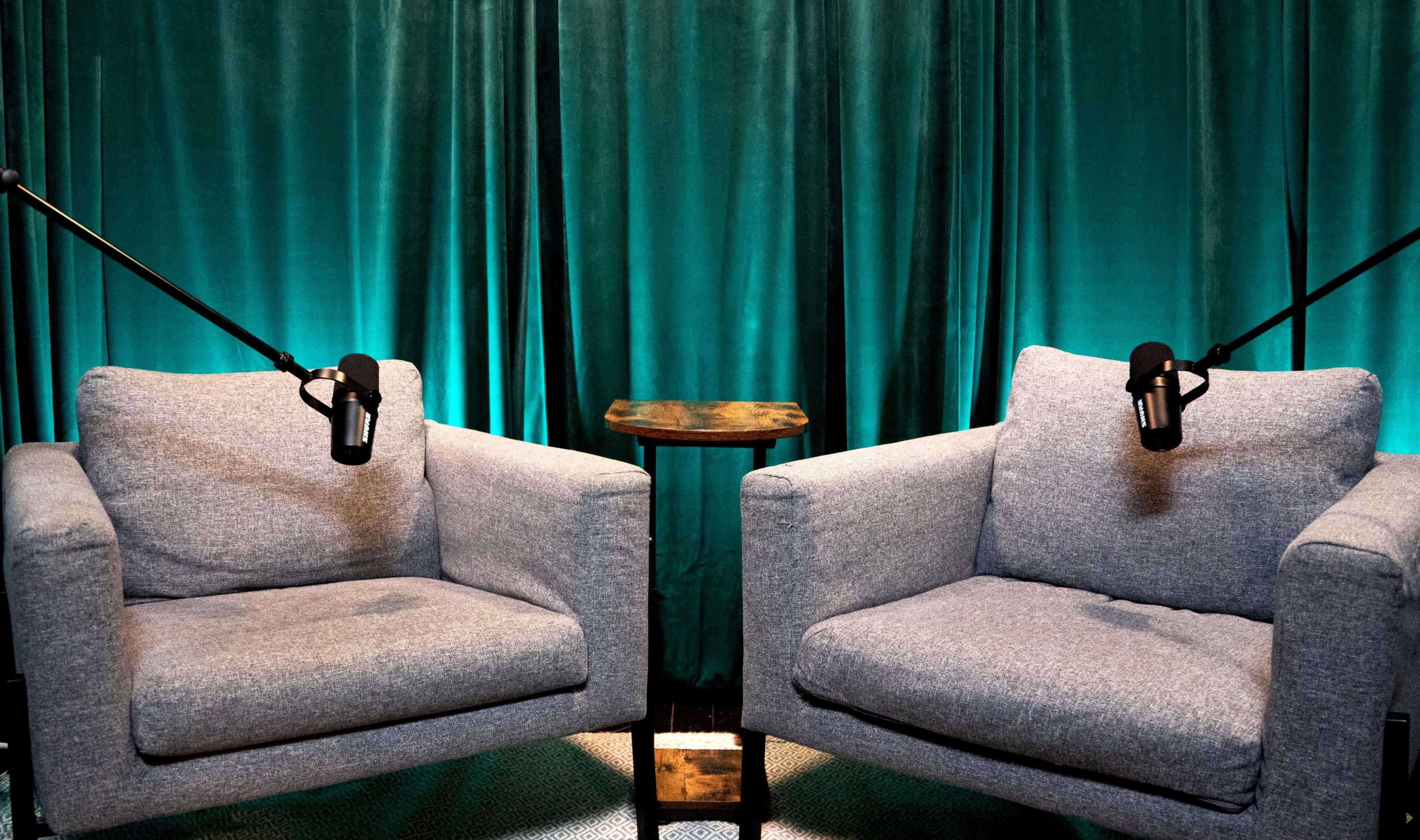 Two gray armchairs with microphones on stands are positioned opposite each other in front of a teal backdrop.