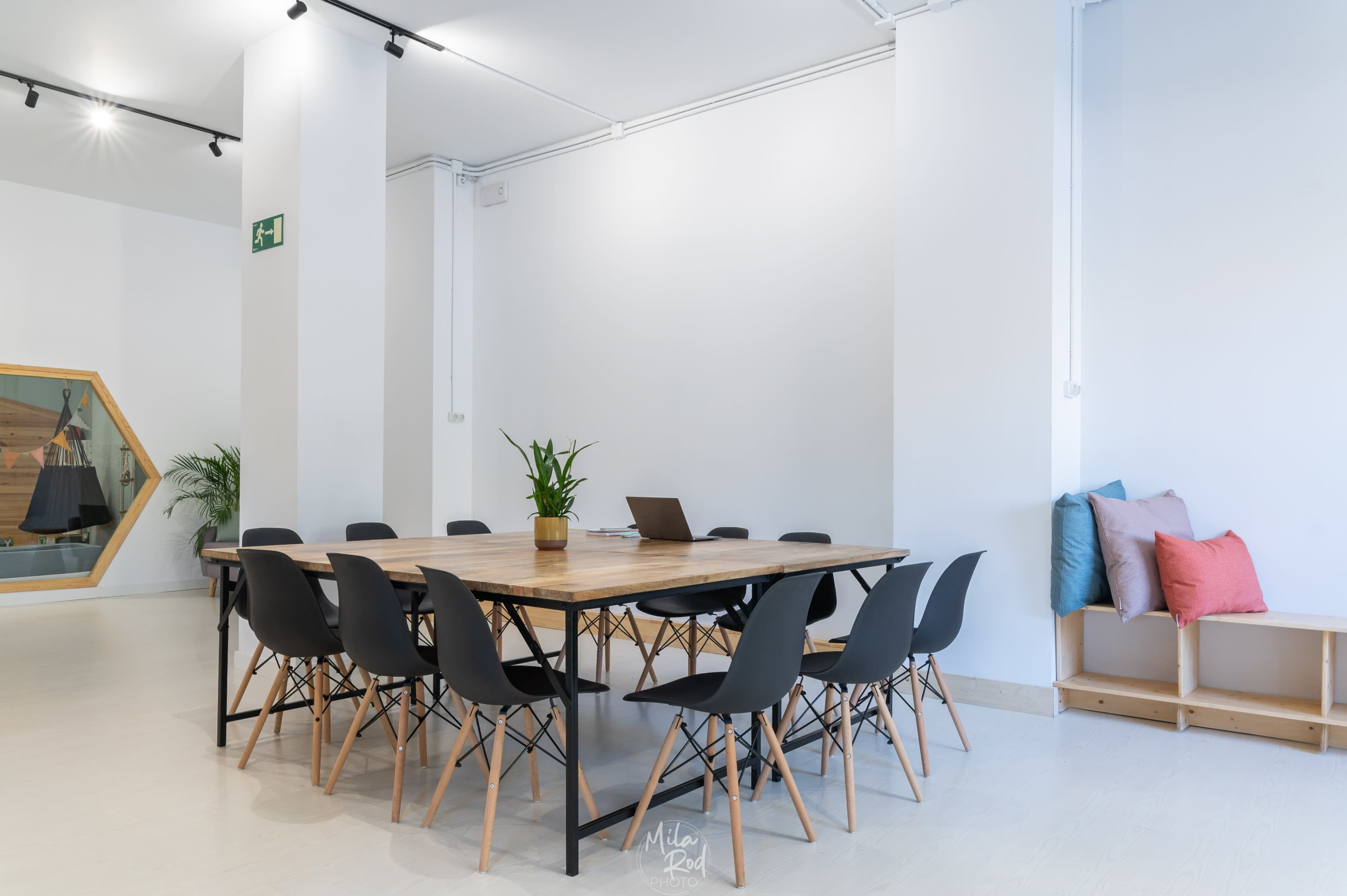 A modern conference room features a large wooden table surrounded by black chairs, with a laptop and a potted plant on the table, in a bright space with minimal decor.
