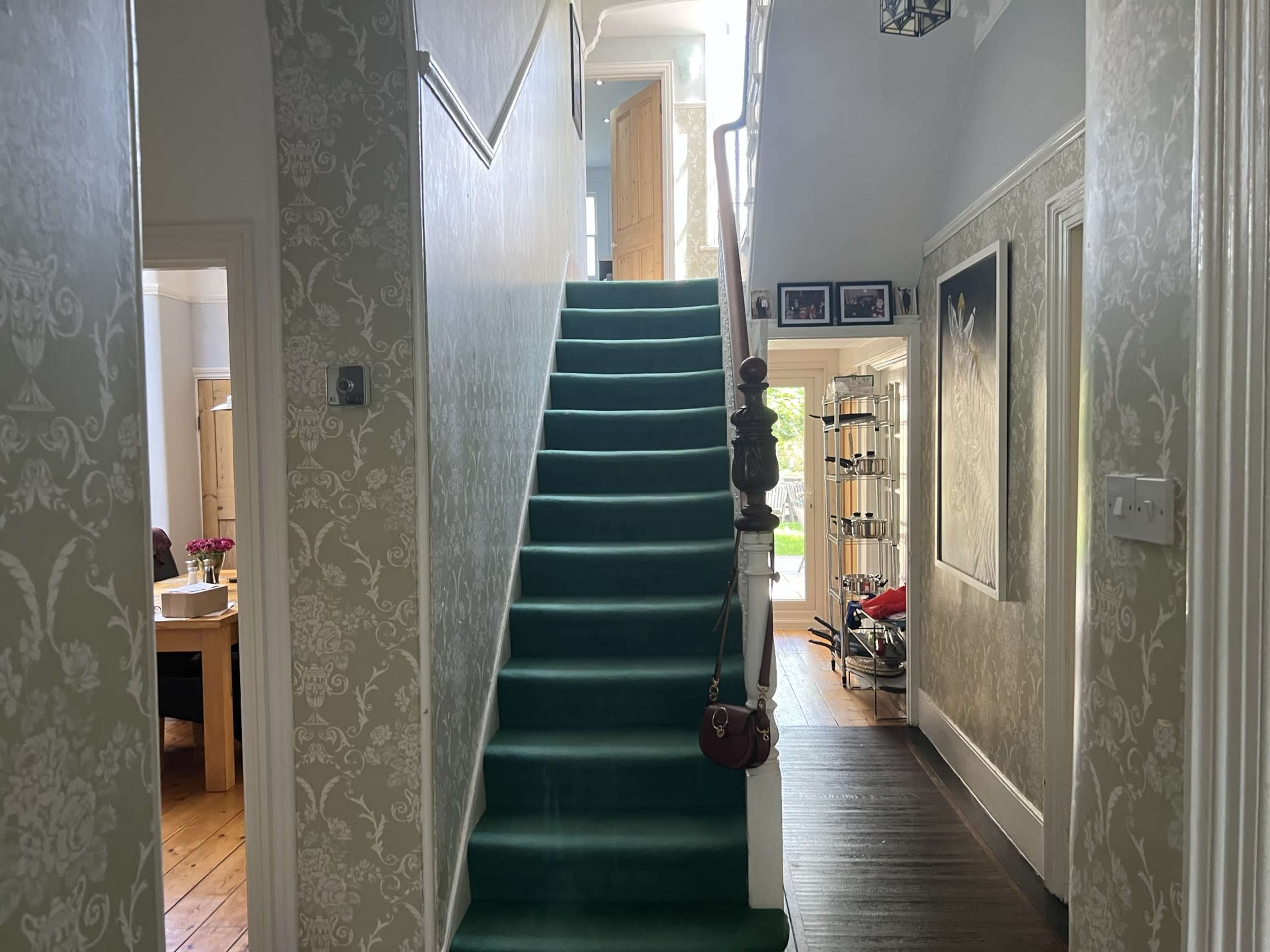 The image shows a staircase with green carpet leading to the upper floor, framed by patterned wallpaper and a wooden floor, with doors and a hallway visible on either side.