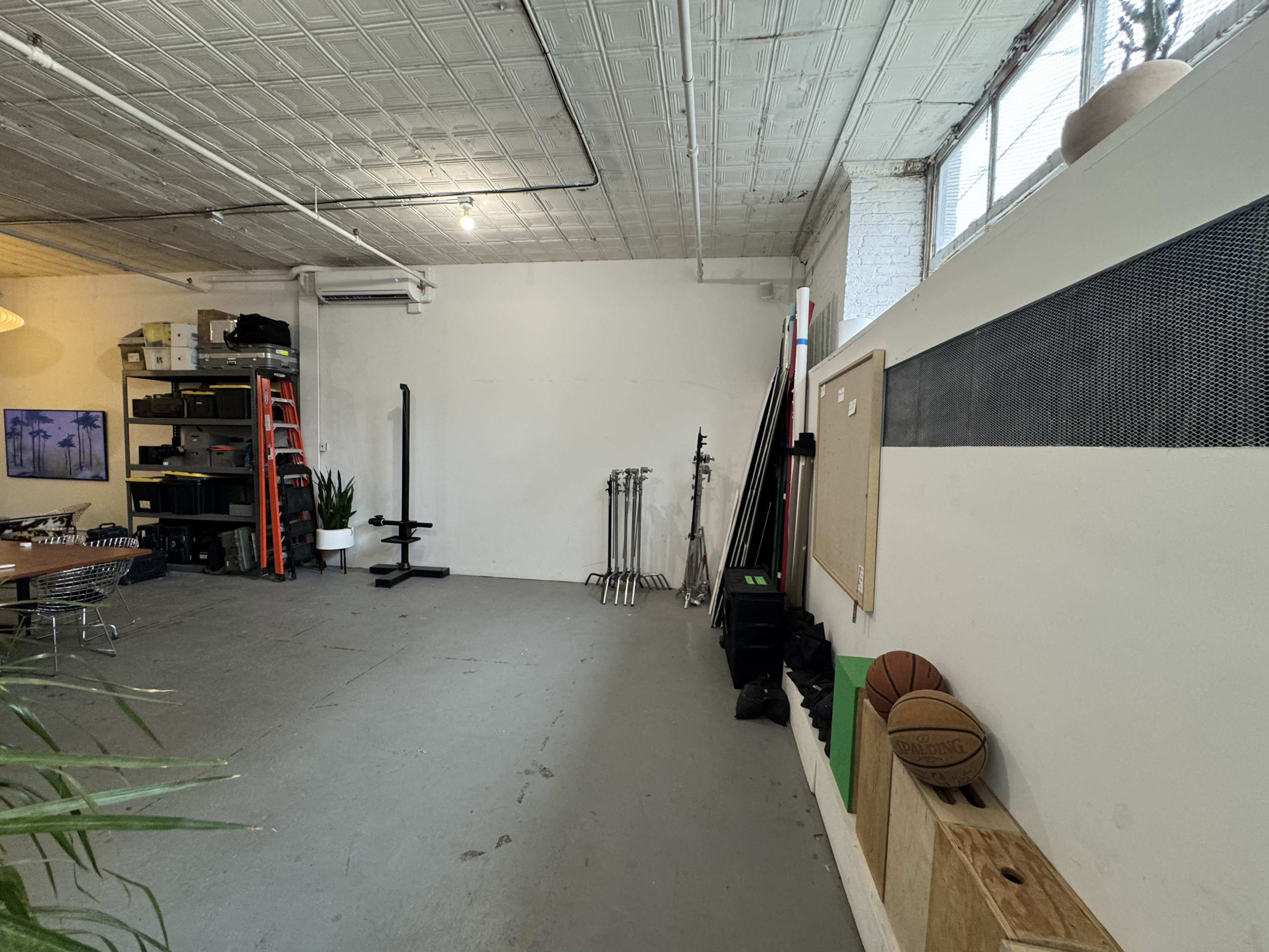 The image shows a spacious garage or studio with a concrete floor, a blank white wall, and various equipment and furniture arranged in the room.