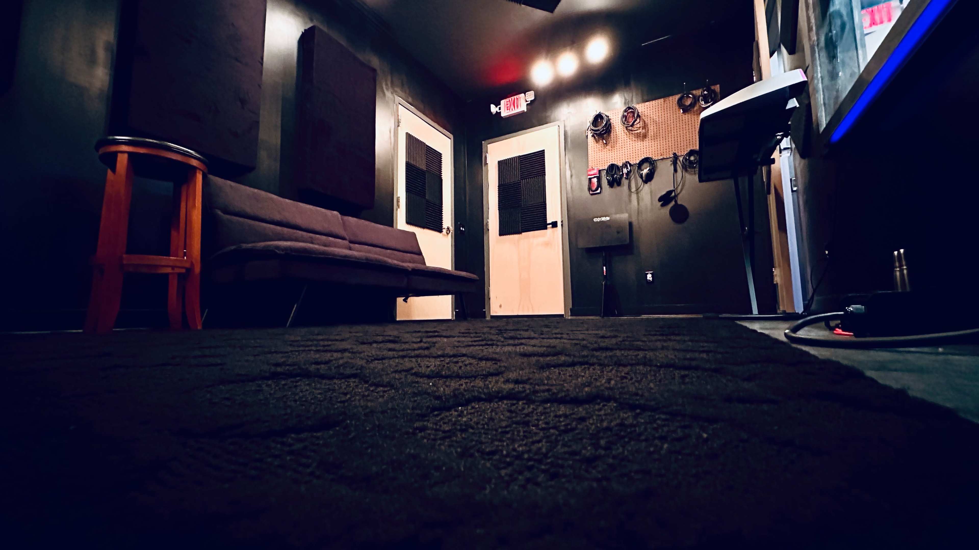 A dimly lit music rehearsal room with a couch, soundproofing panels on the walls, and various musical equipment mounted nearby.