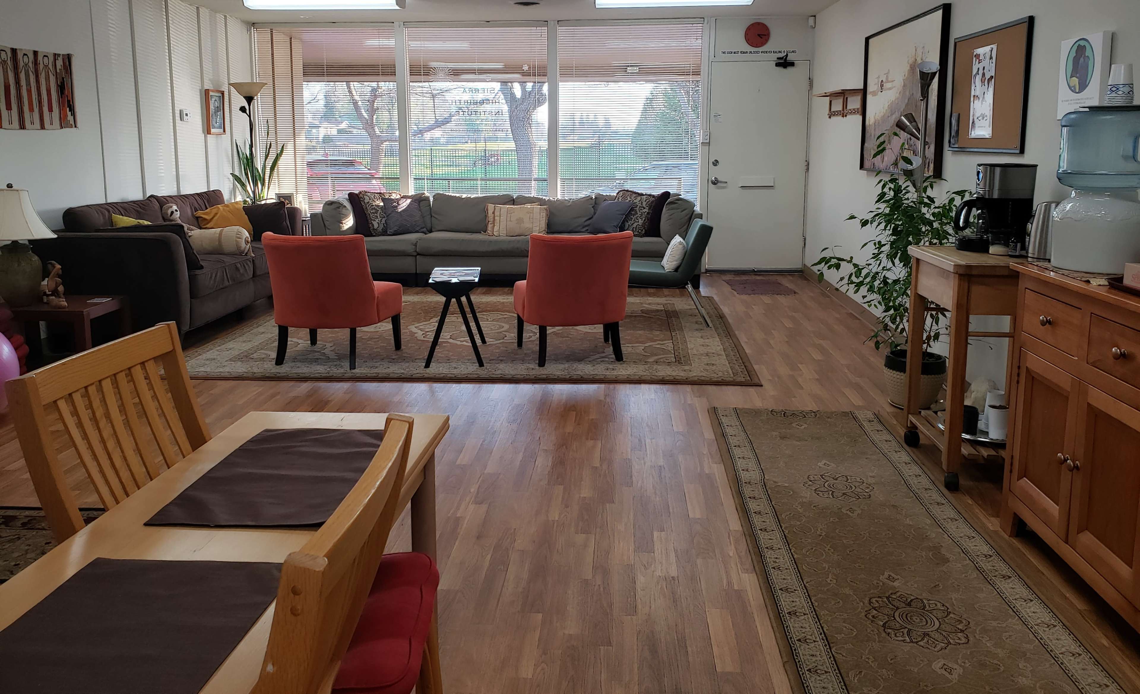 A cozy living space featuring a gray couch, two orange chairs, a wooden dining table, and a water dispenser against a backdrop of large windows.
