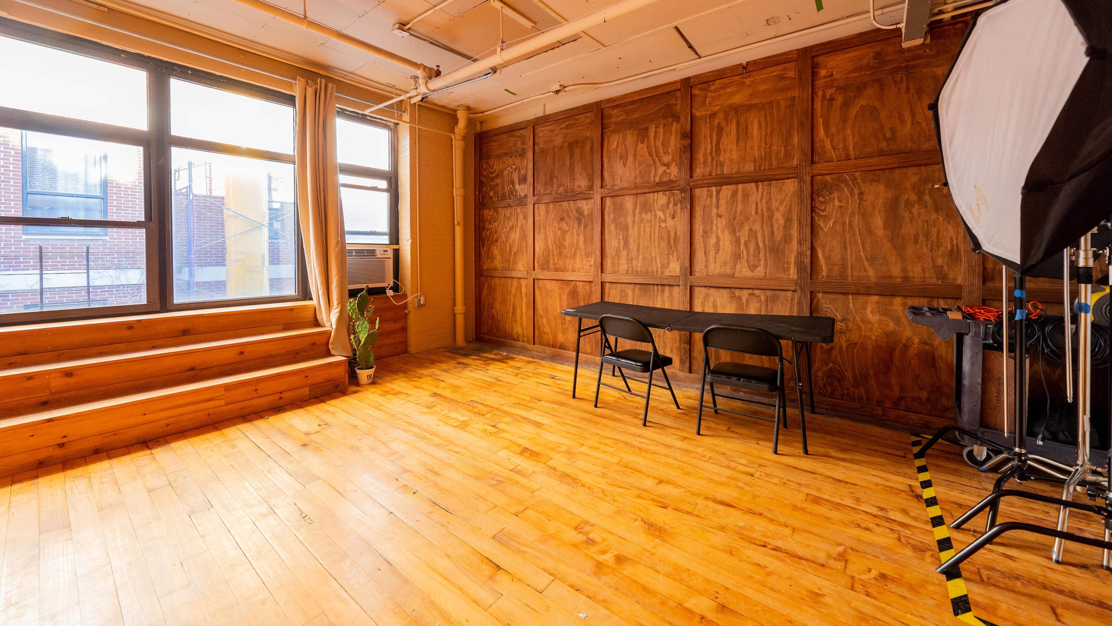 A spacious, wooden-floored room with large windows, a table and chairs, and a minimalistic setup for lighting equipment.