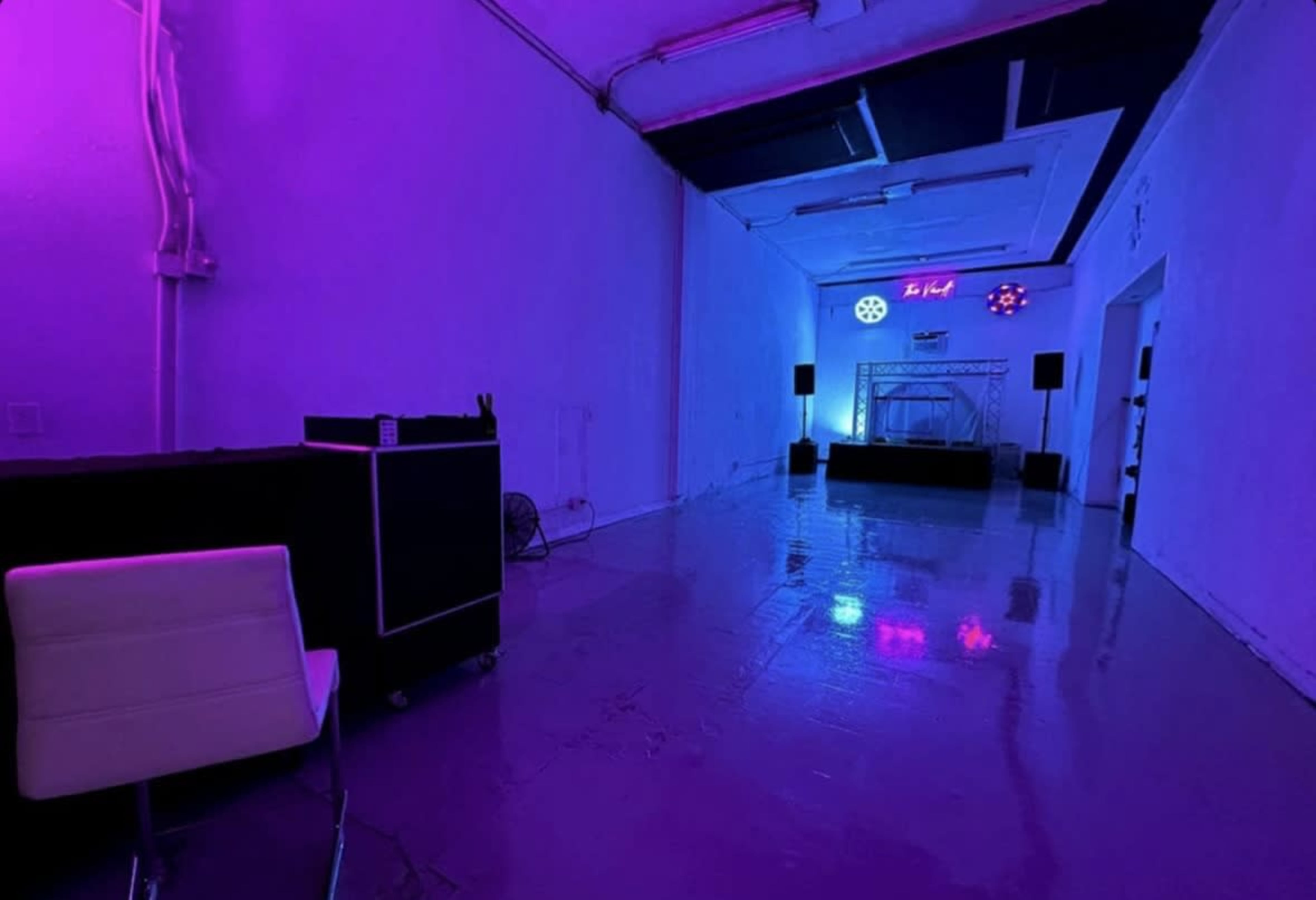 The image shows a spacious room with a reflective floor, illuminated by purple and blue lights, featuring a stage at one end and audio equipment set up along the sides.