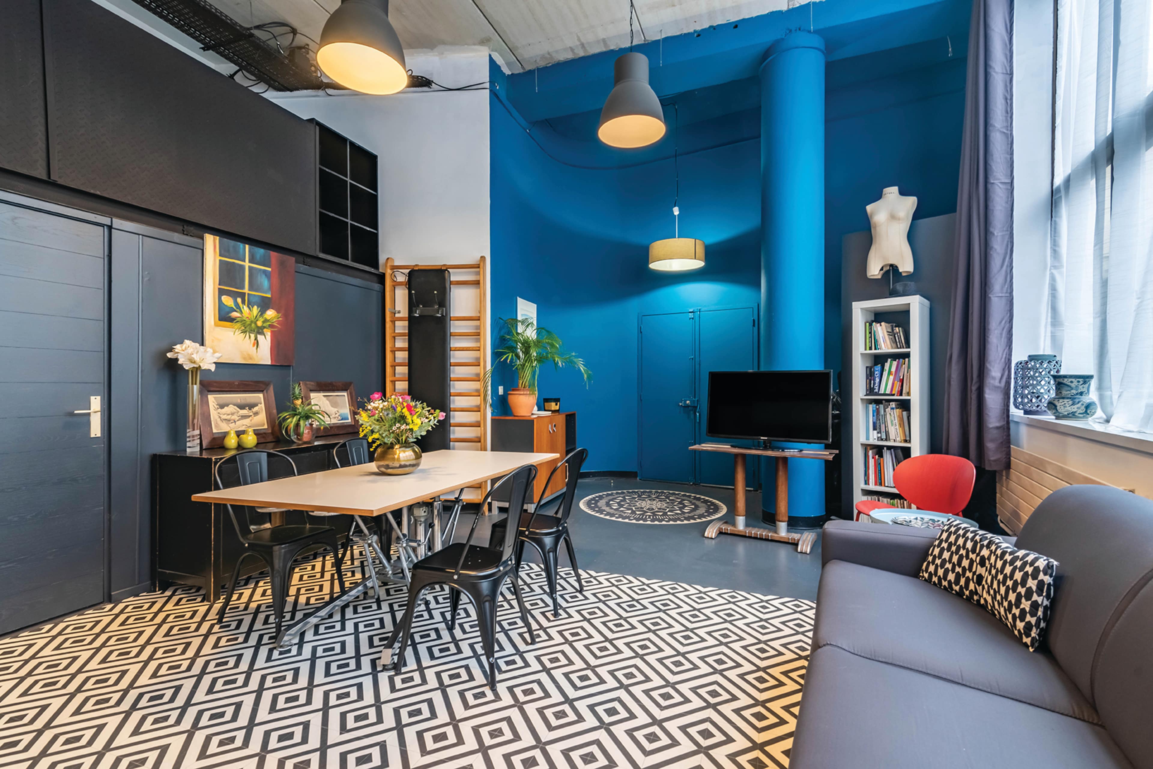 A modern, open living space featuring a dining table, a sofa, a television, and a geometric patterned floor, with a blue wall and various decorative items.