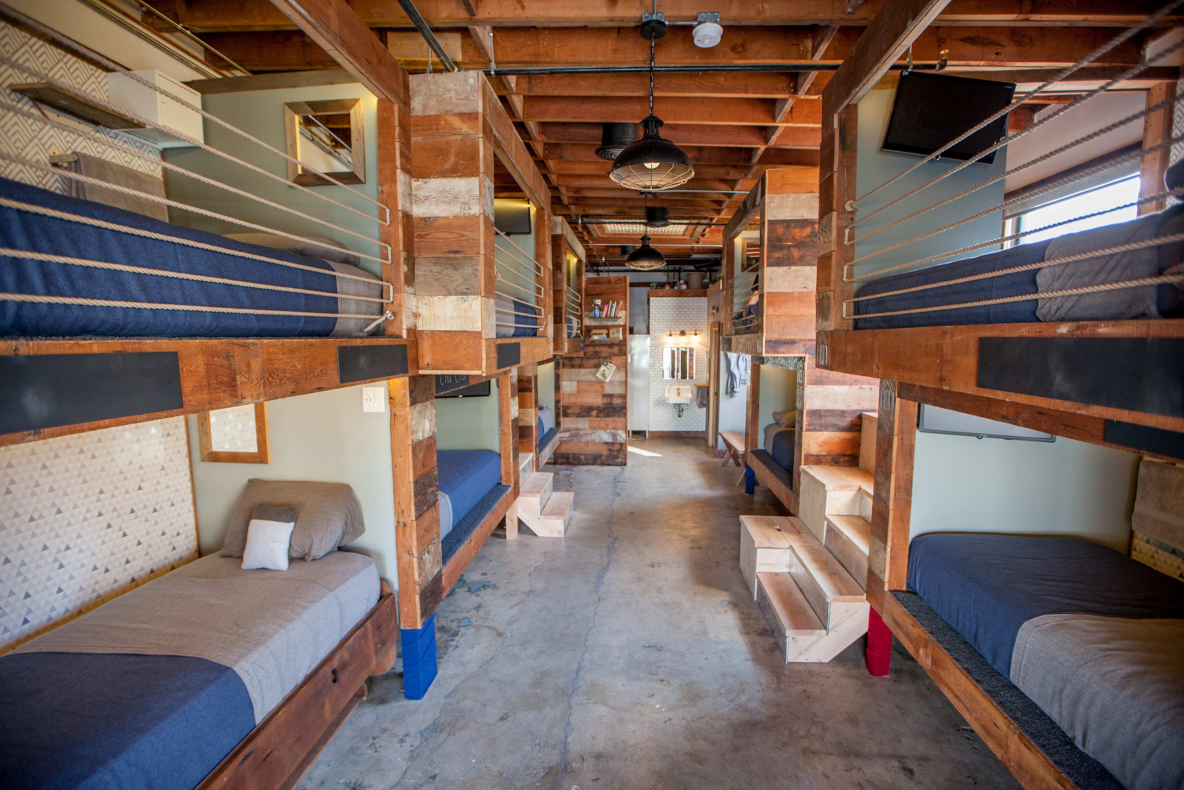 The image shows a spacious dormitory with multiple bunk beds arranged in a well-lit room featuring wooden beams and a communal area in the background.
