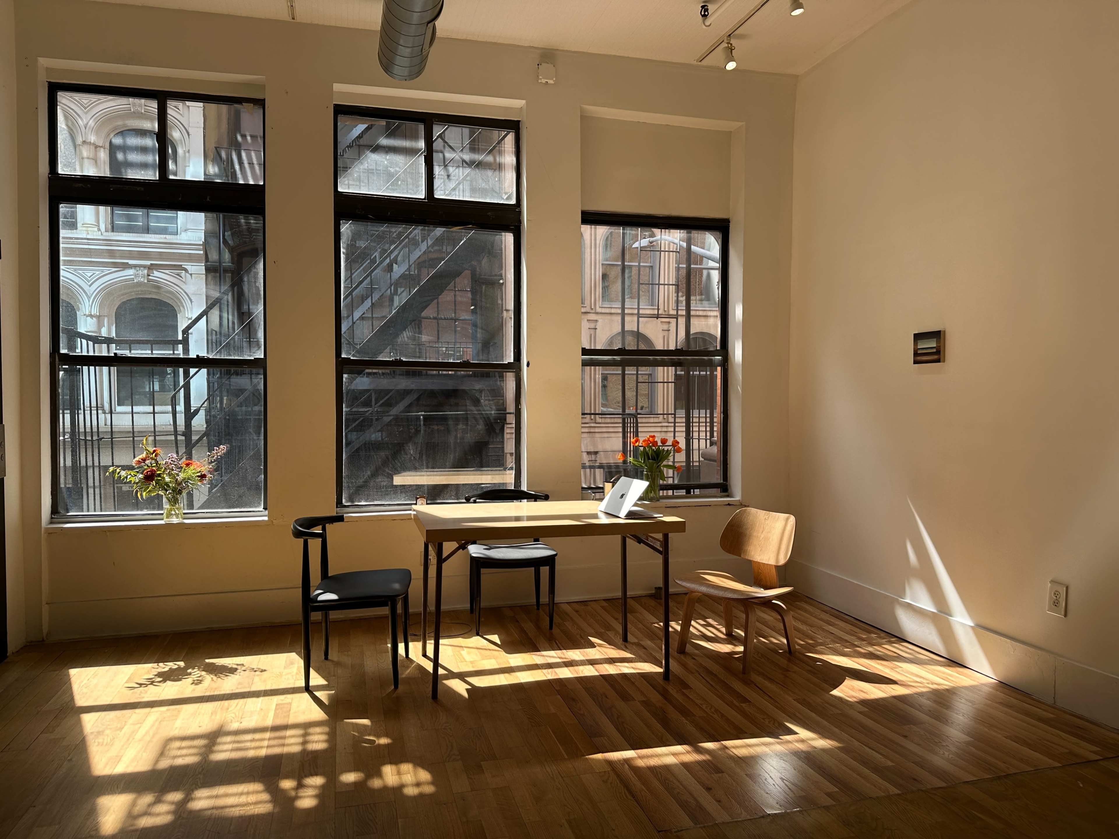 A small room features a table with a laptop and two chairs, illuminated by sunlight filtering through large windows.