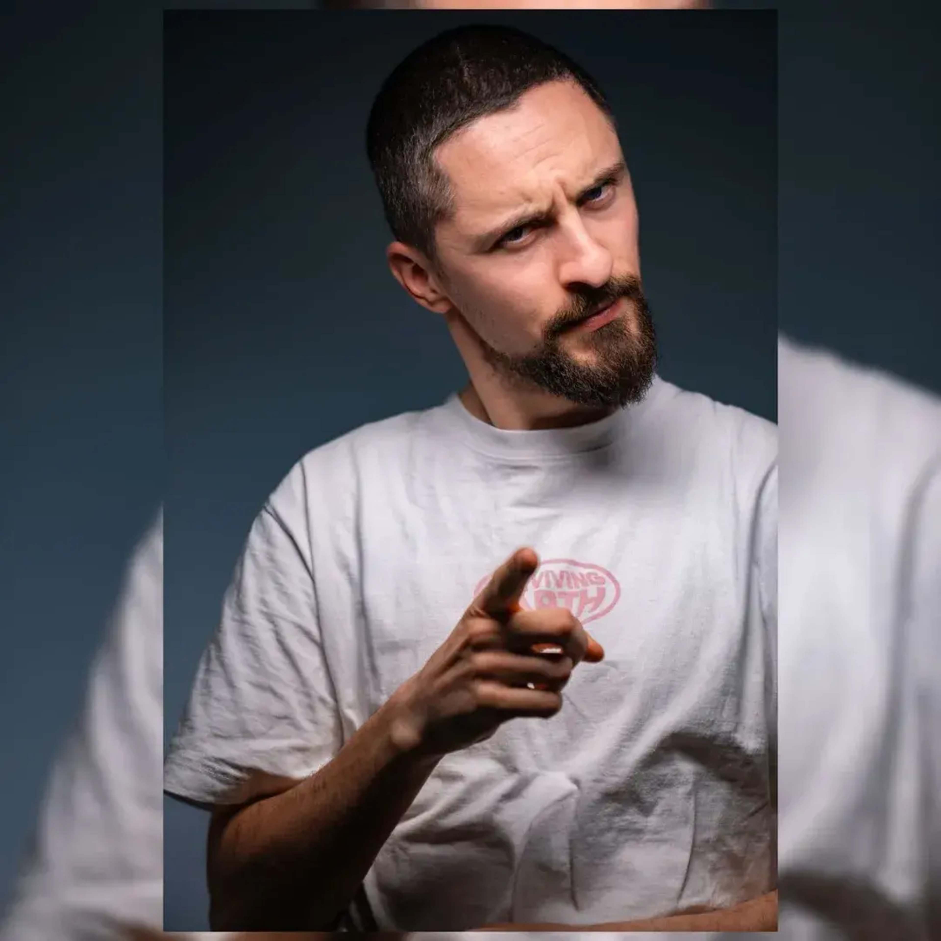 A man with a beard poses with a serious expression, pointing his finger while wearing a plain white t-shirt.