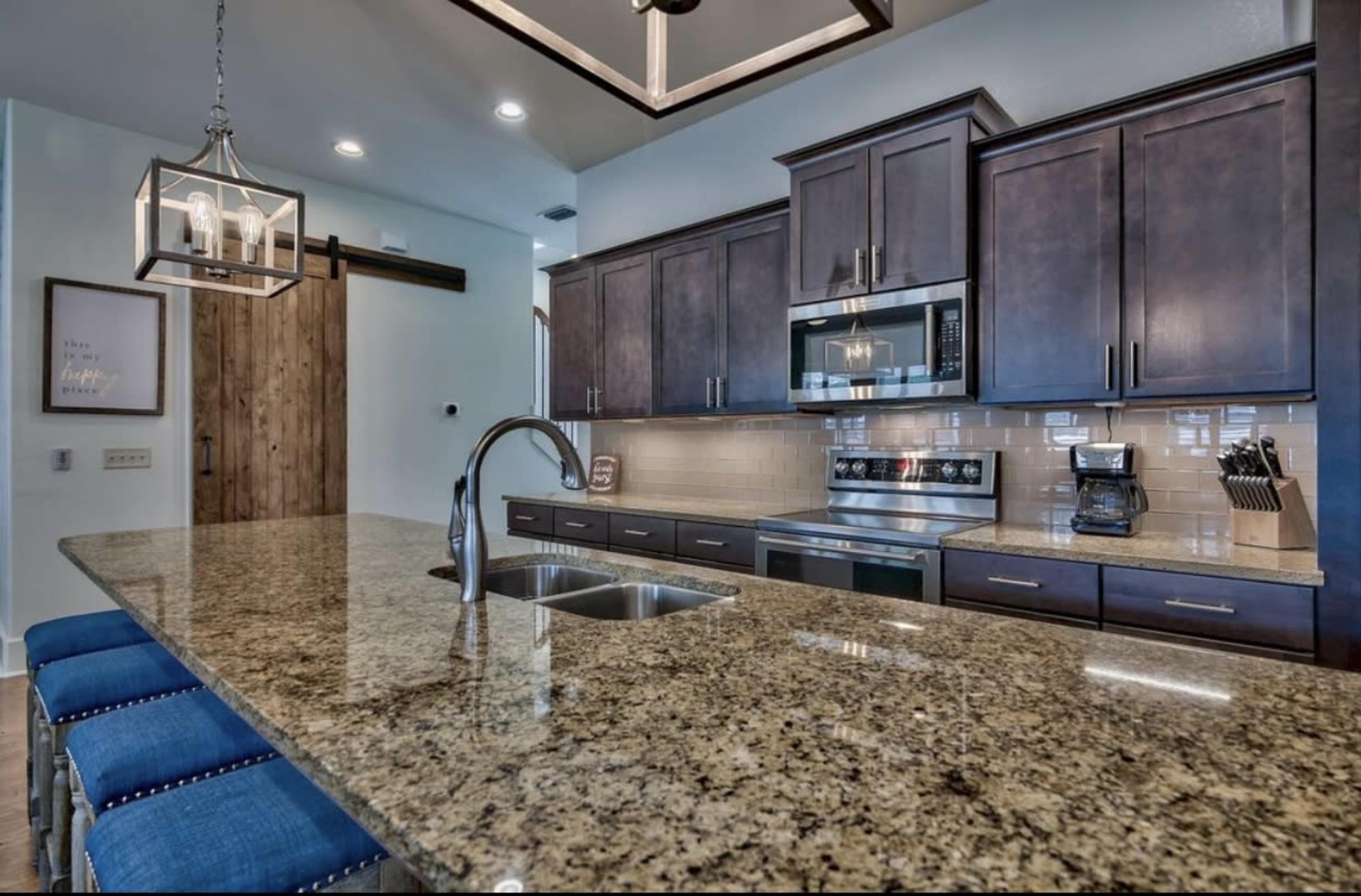 A modern kitchen with dark wooden cabinets, stainless steel appliances, a granite countertop with a double sink, and blue upholstered bar stools.