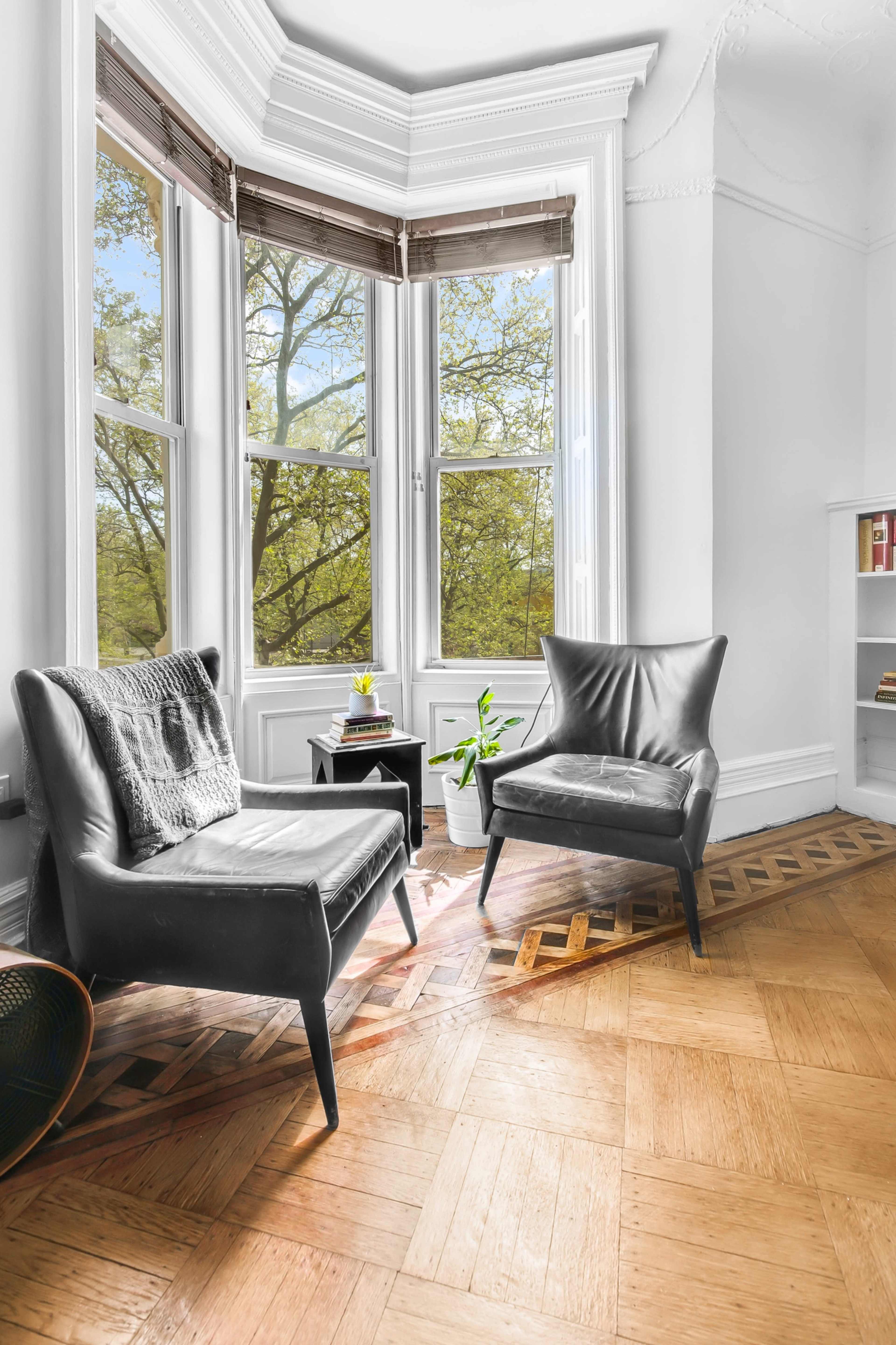 The image shows a bright corner of a room with two black armchairs positioned near large windows overlooking trees outside.