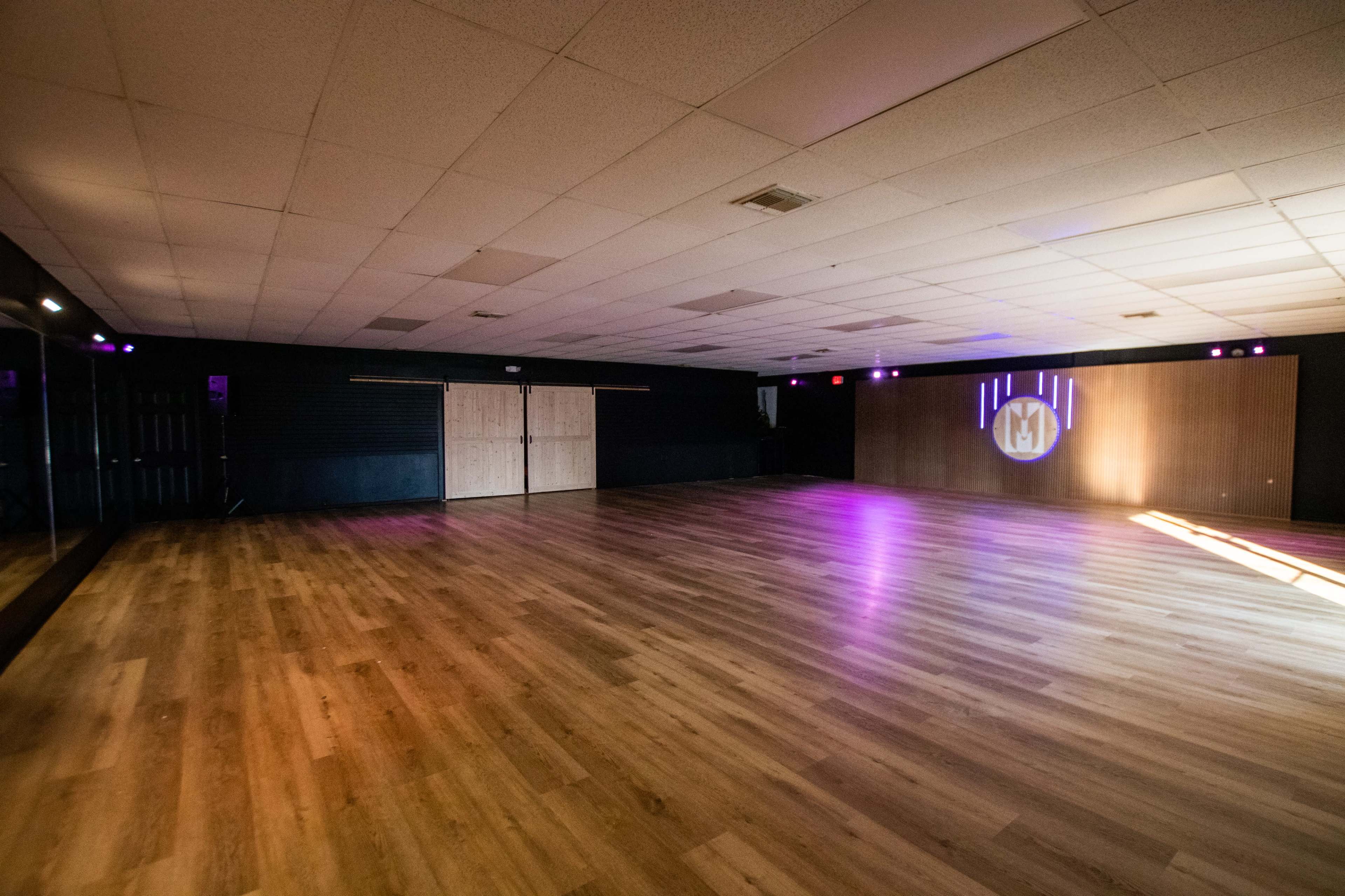The image shows an empty dance studio with a wooden floor, mirrored walls, and soft purple lighting.