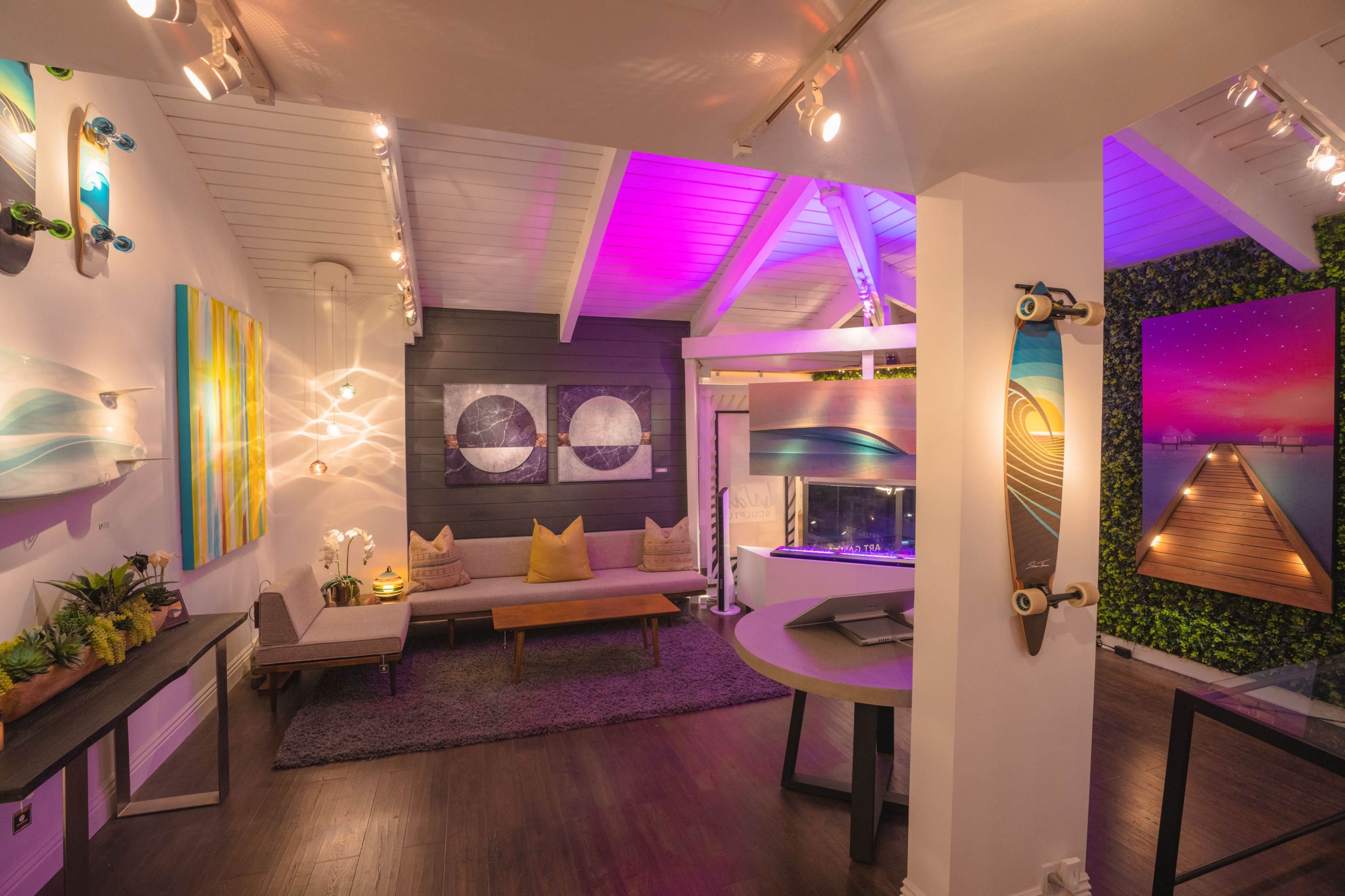A modern, brightly lit art gallery with a mix of painted canvases and surfboard art displayed on the walls, along with a cozy seating area.