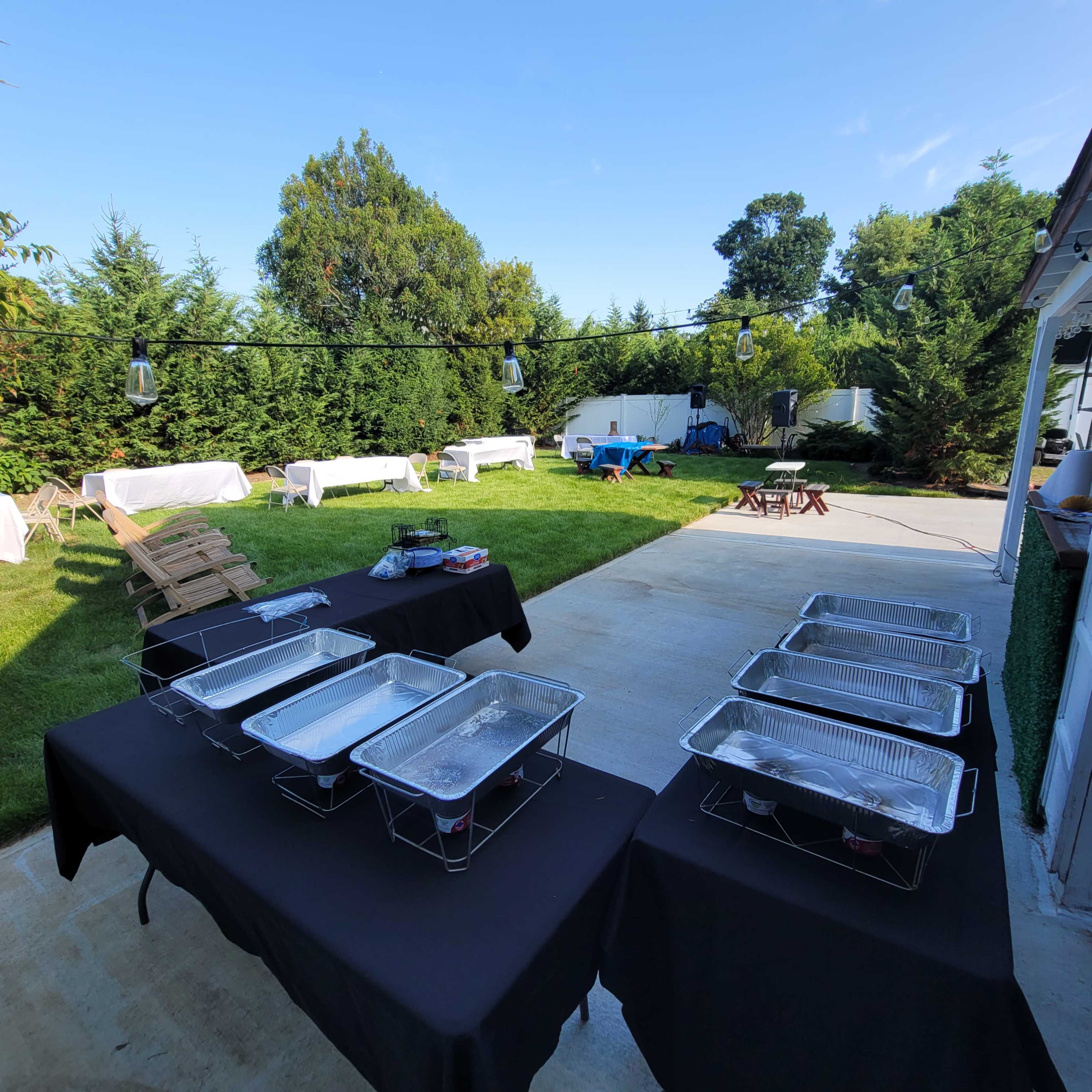 A backyard is set up for a gathering, featuring several tables with food trays, outdoor seating, and green foliage surrounding the area.
