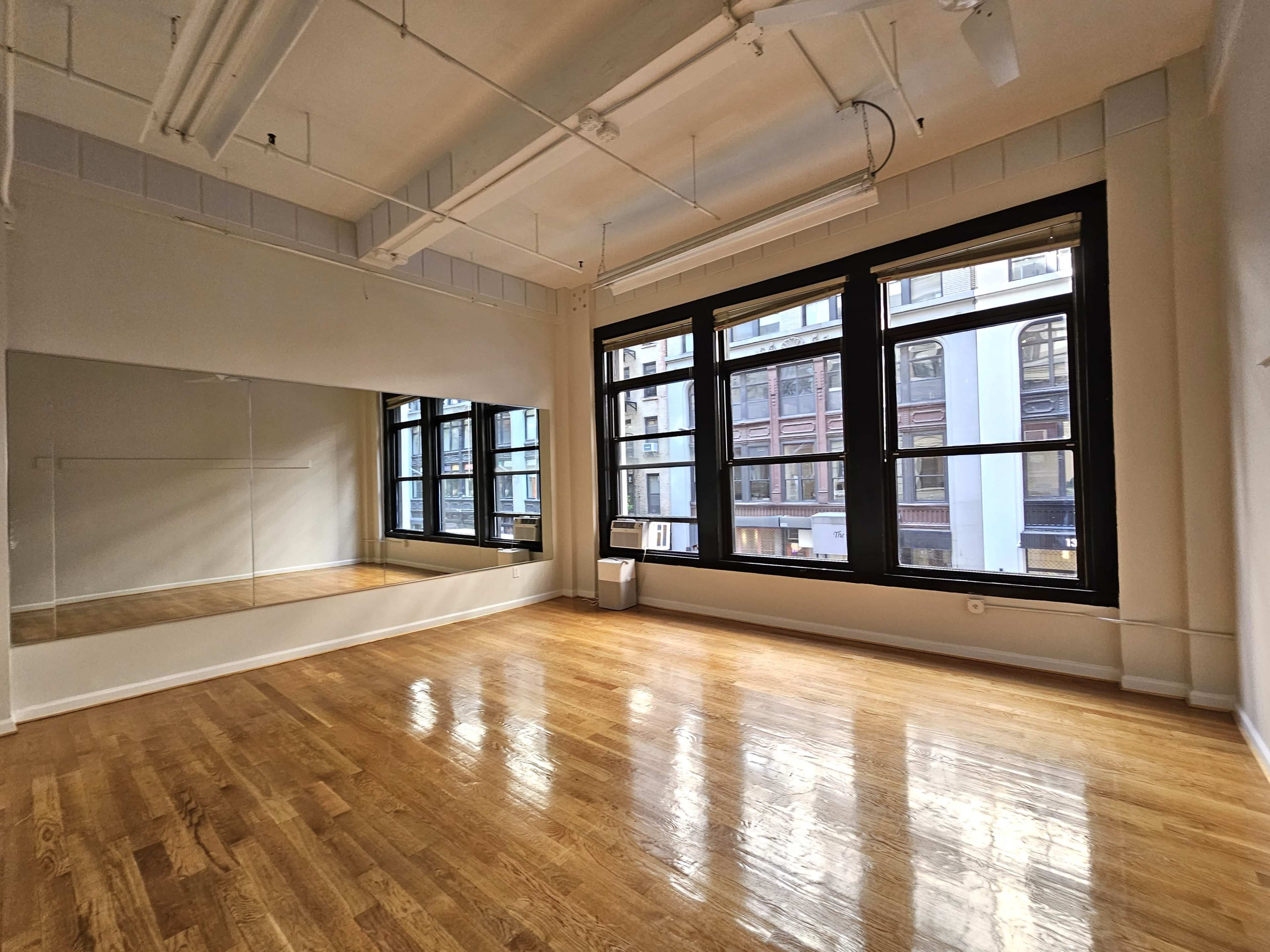 An empty room with wooden floors, large windows, and a mirrored wall reflecting the windows.