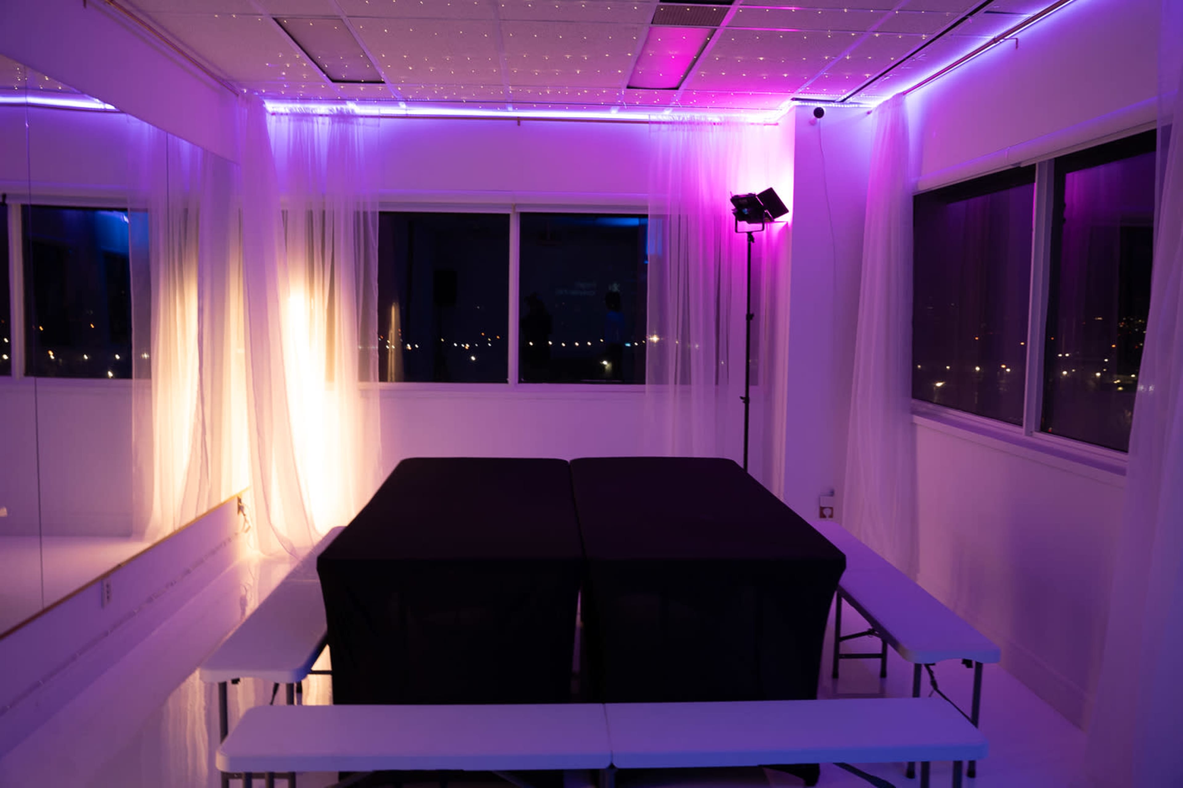 The image shows a brightly lit event space with two black tables set against a backdrop of soft drapes and purple lighting.
