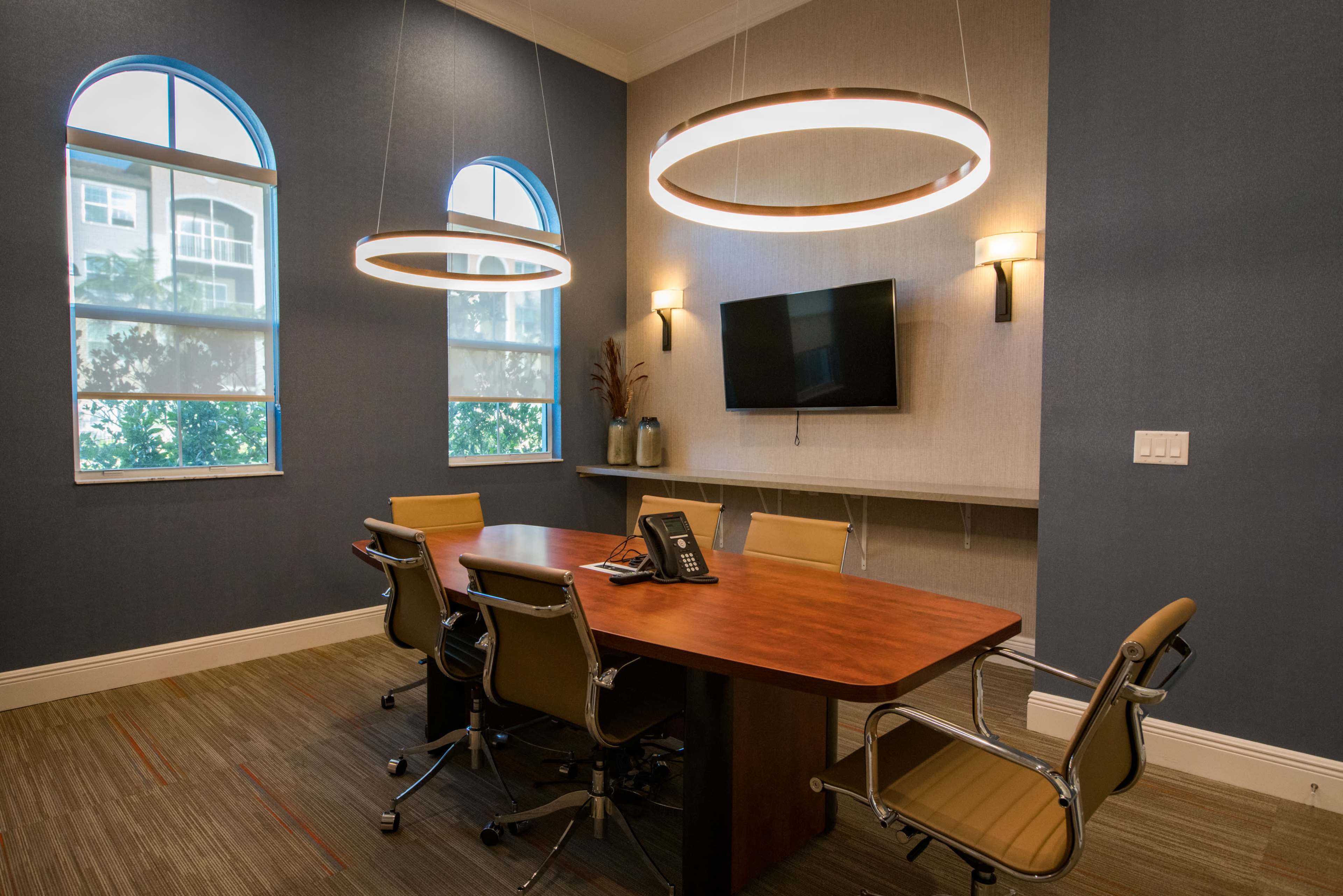 A conference room features a wooden table surrounded by six chairs, with large light fixtures above and a wall-mounted television.
