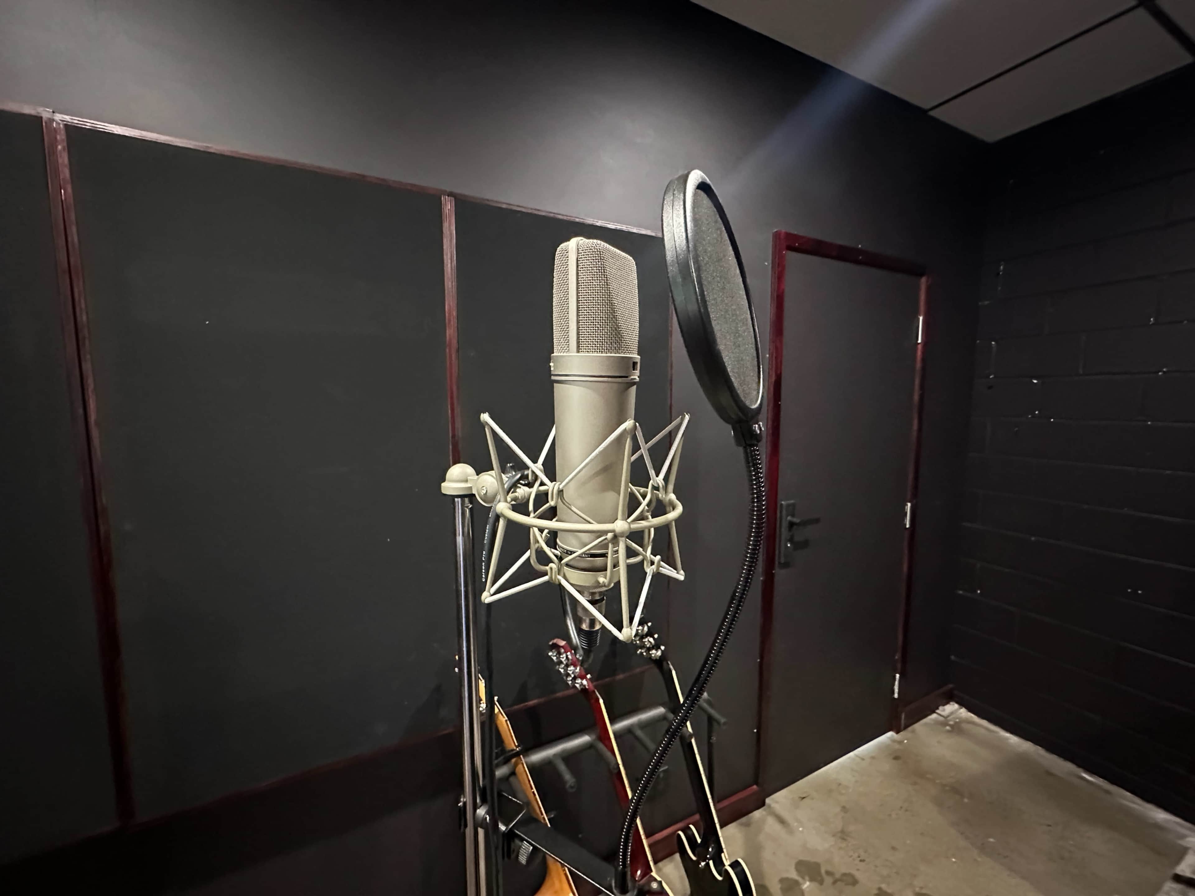A microphone stand with a large condenser microphone in a recording studio, alongside a guitar leaning against the wall.