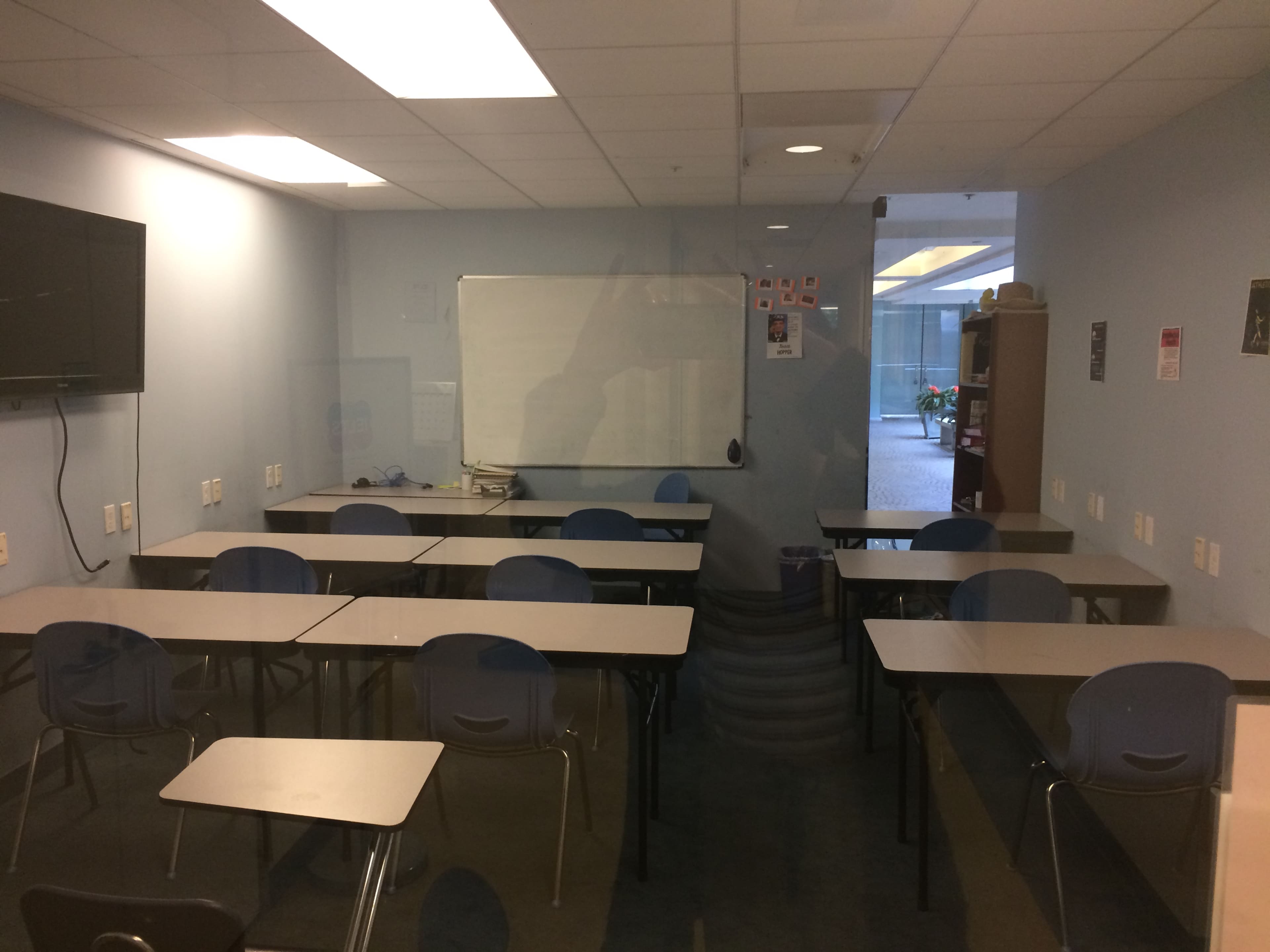 A classroom is depicted with rows of empty desks facing a whiteboard and a television mounted on the wall.