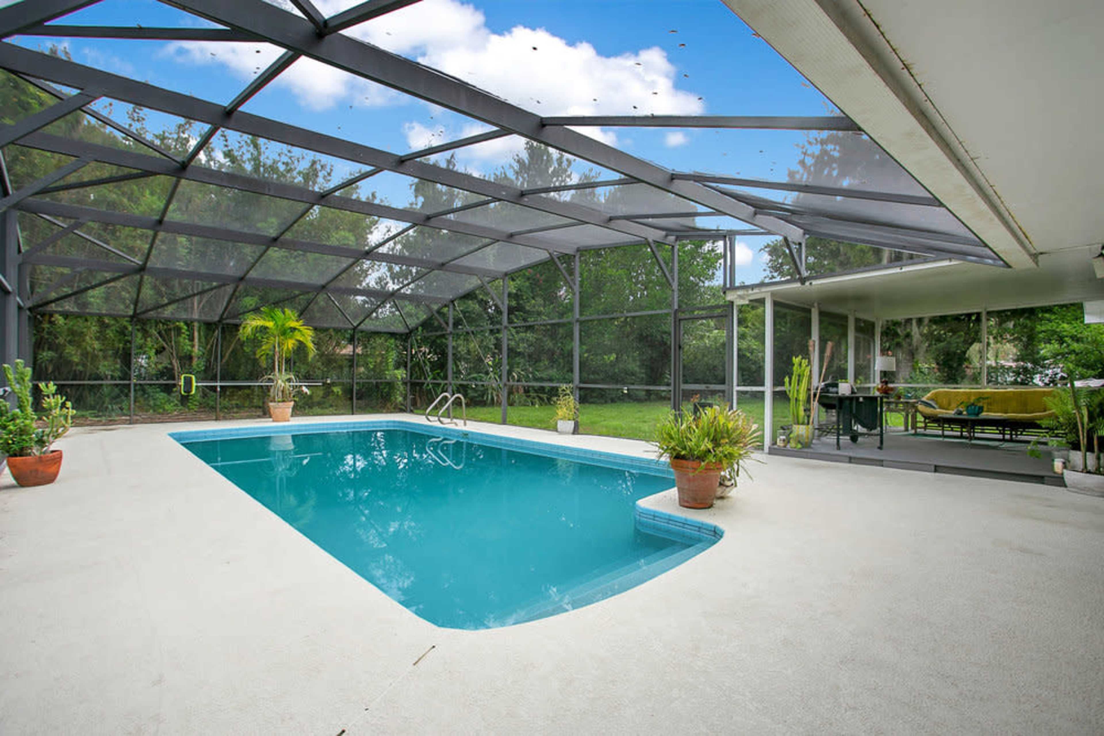 The image shows an outdoor swimming pool enclosed by a screen, surrounded by potted plants and a shaded seating area.