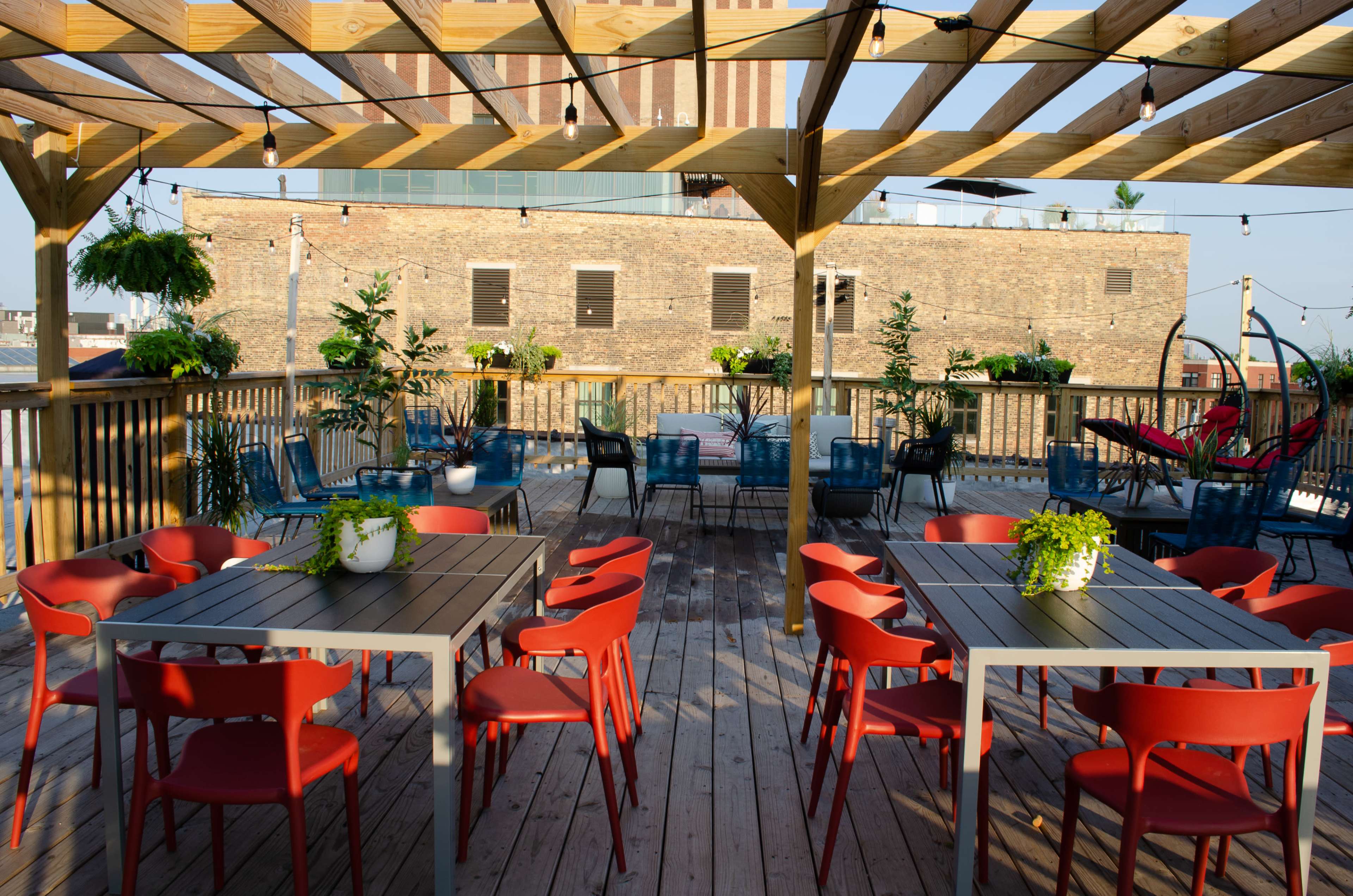 A rooftop deck with wooden flooring and several tables surrounded by potted plants and red chairs.