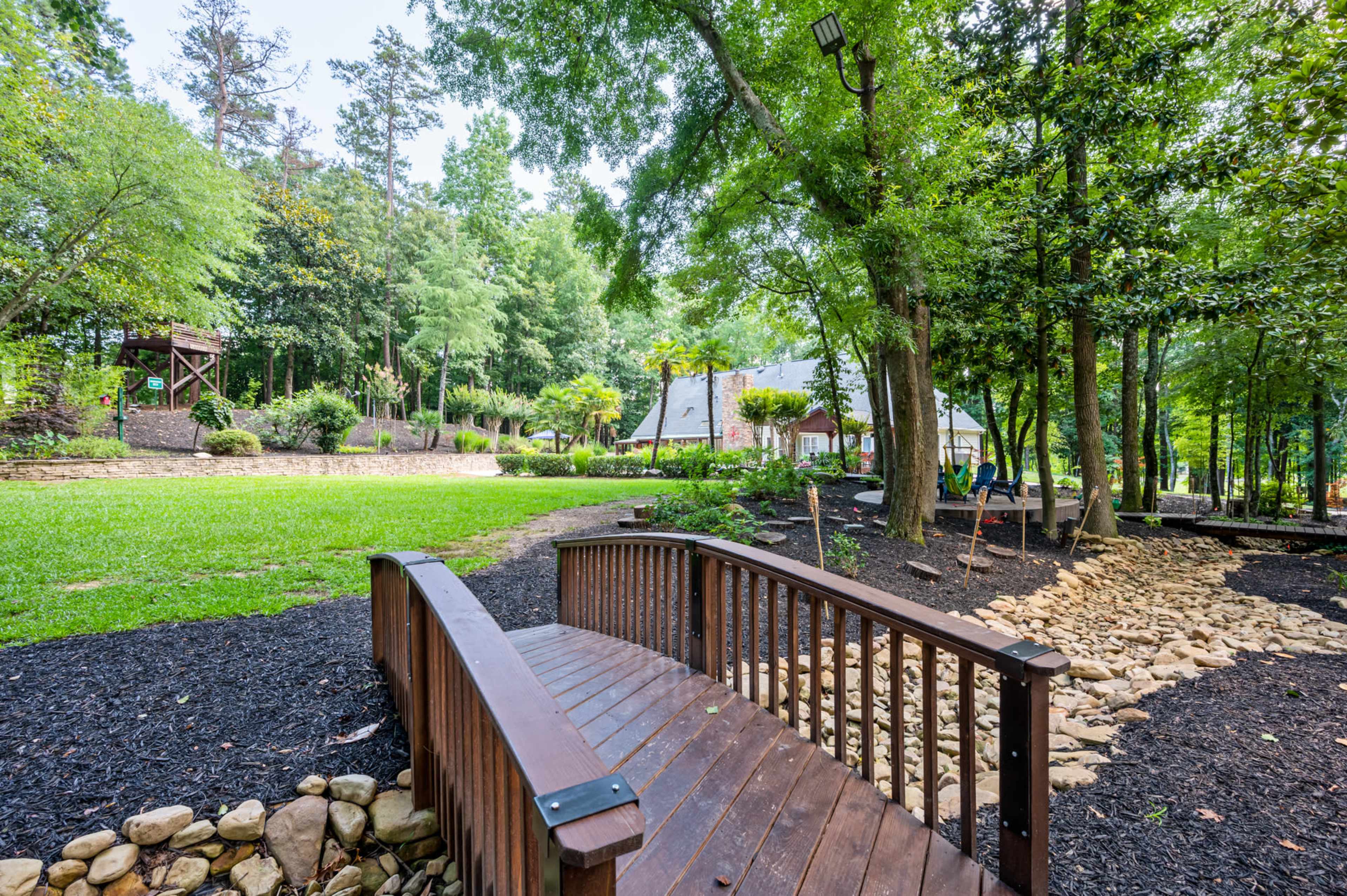 A wooden bridge arching over a path made of stones in a landscaped park surrounded by trees and a grassy area.