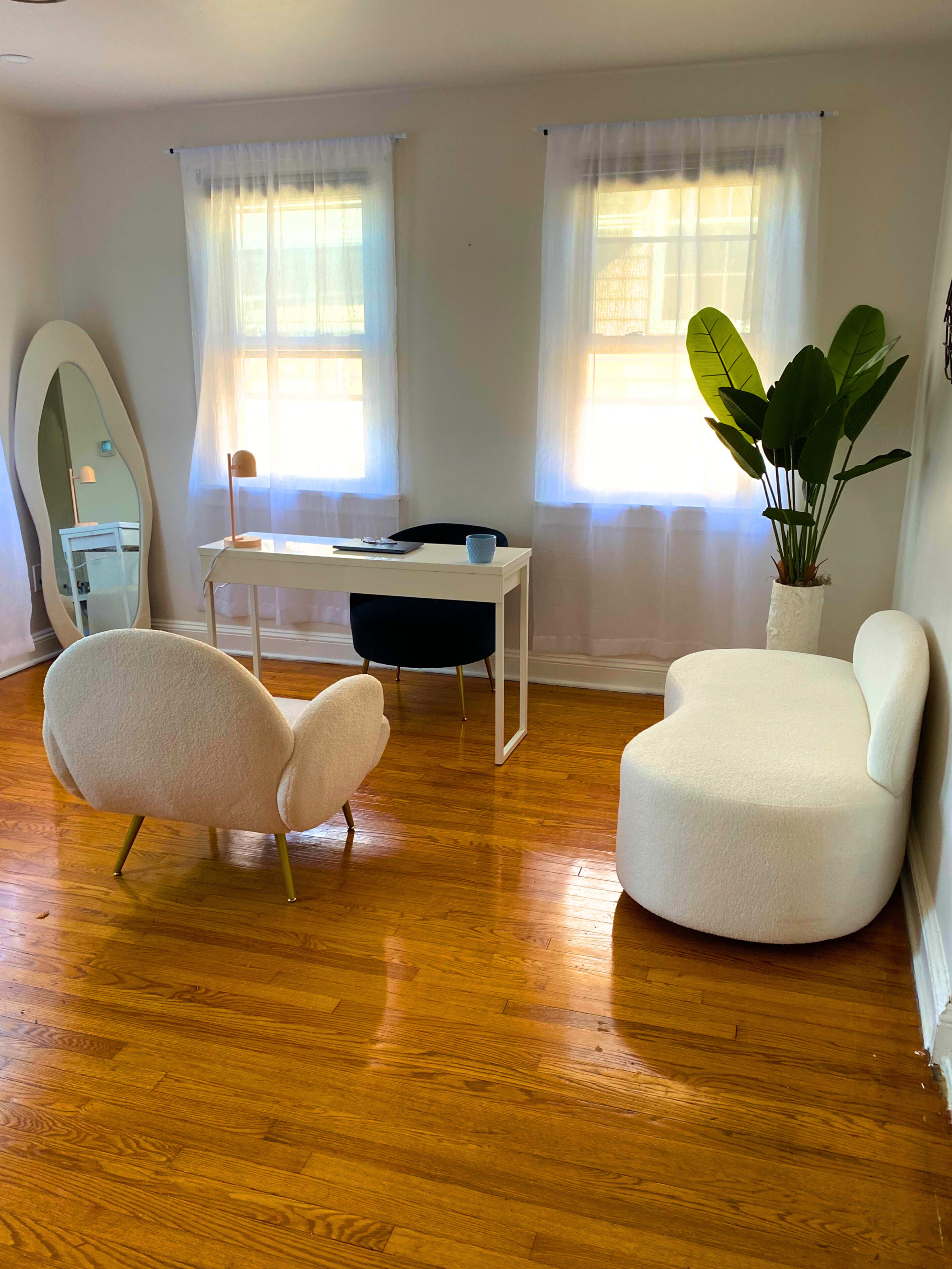 A bright room featuring a white desk with a chair, two modern chairs, a large mirror, and a potted plant.