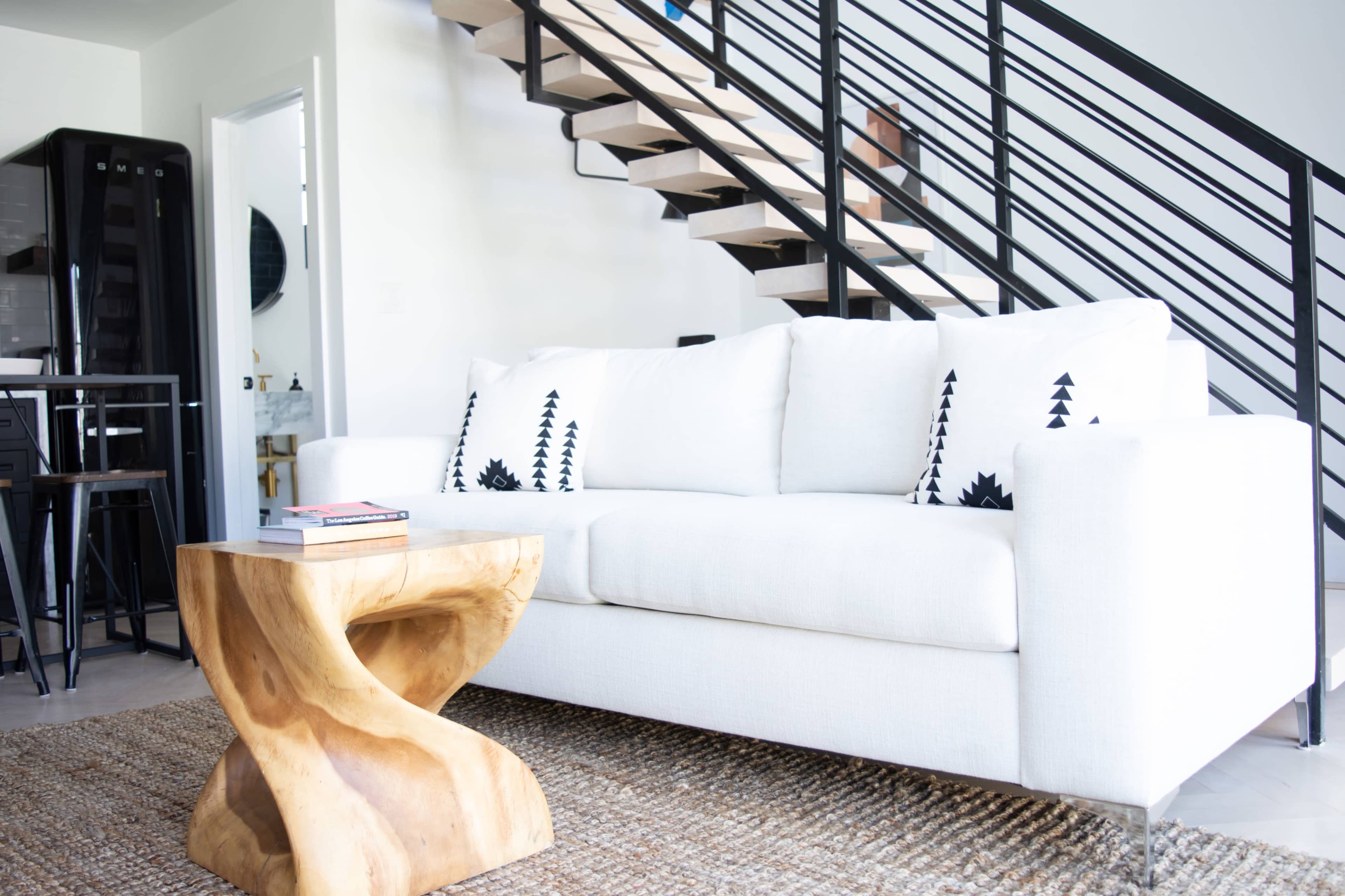 A modern living space featuring a white sofa with patterned throw pillows, a wooden sculpture table, and a staircase with a black railing in the background.