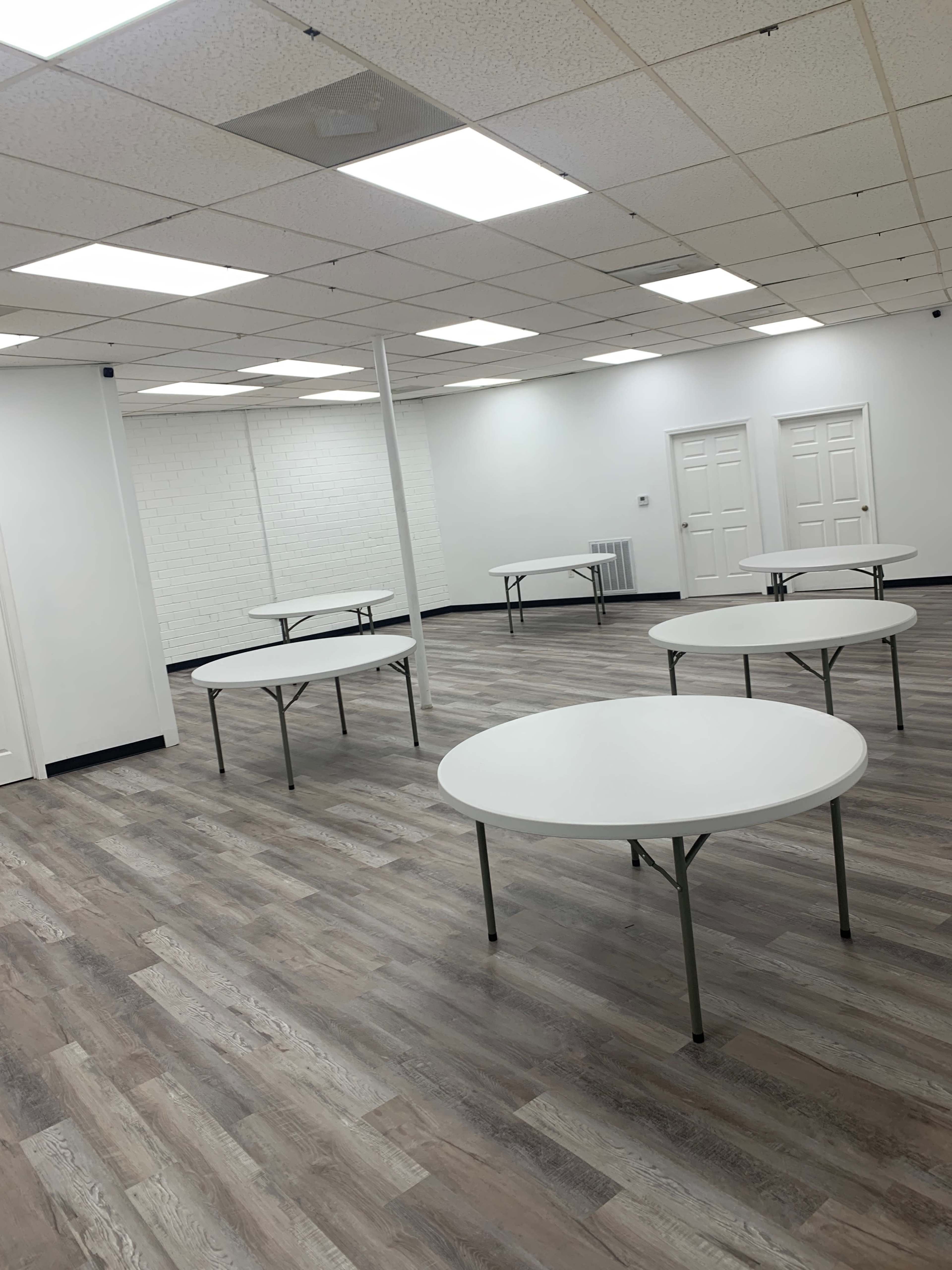 The image shows a spacious, well-lit room with several round tables arranged in a grid pattern on a wooden floor.