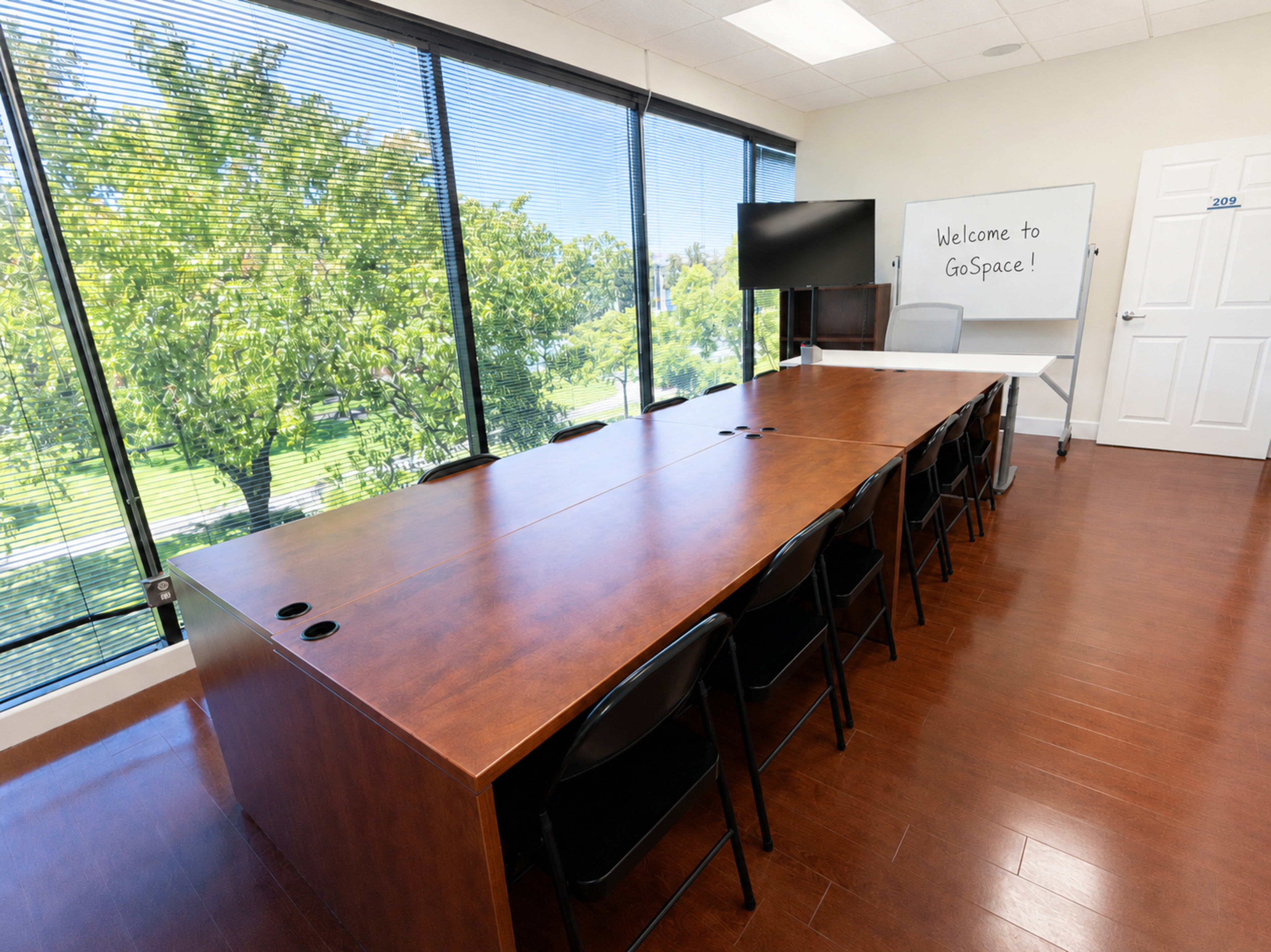The image shows a modern conference room with a long wooden table, black chairs, large windows overlooking green trees, and a whiteboard with a welcome message.