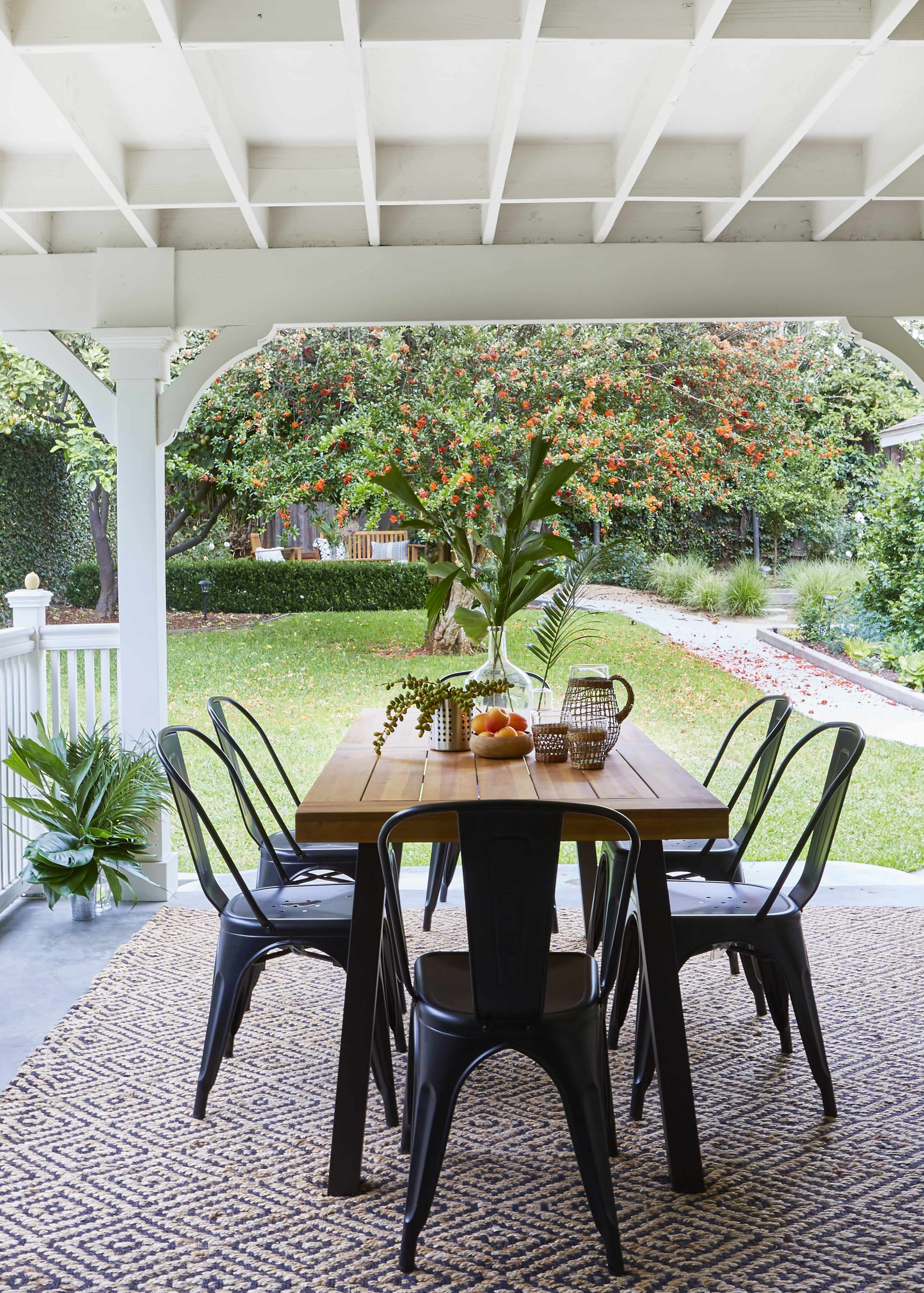 A wooden dining table with black chairs is set under a covered porch, surrounded by greenery and a garden with a flowering tree in the background.